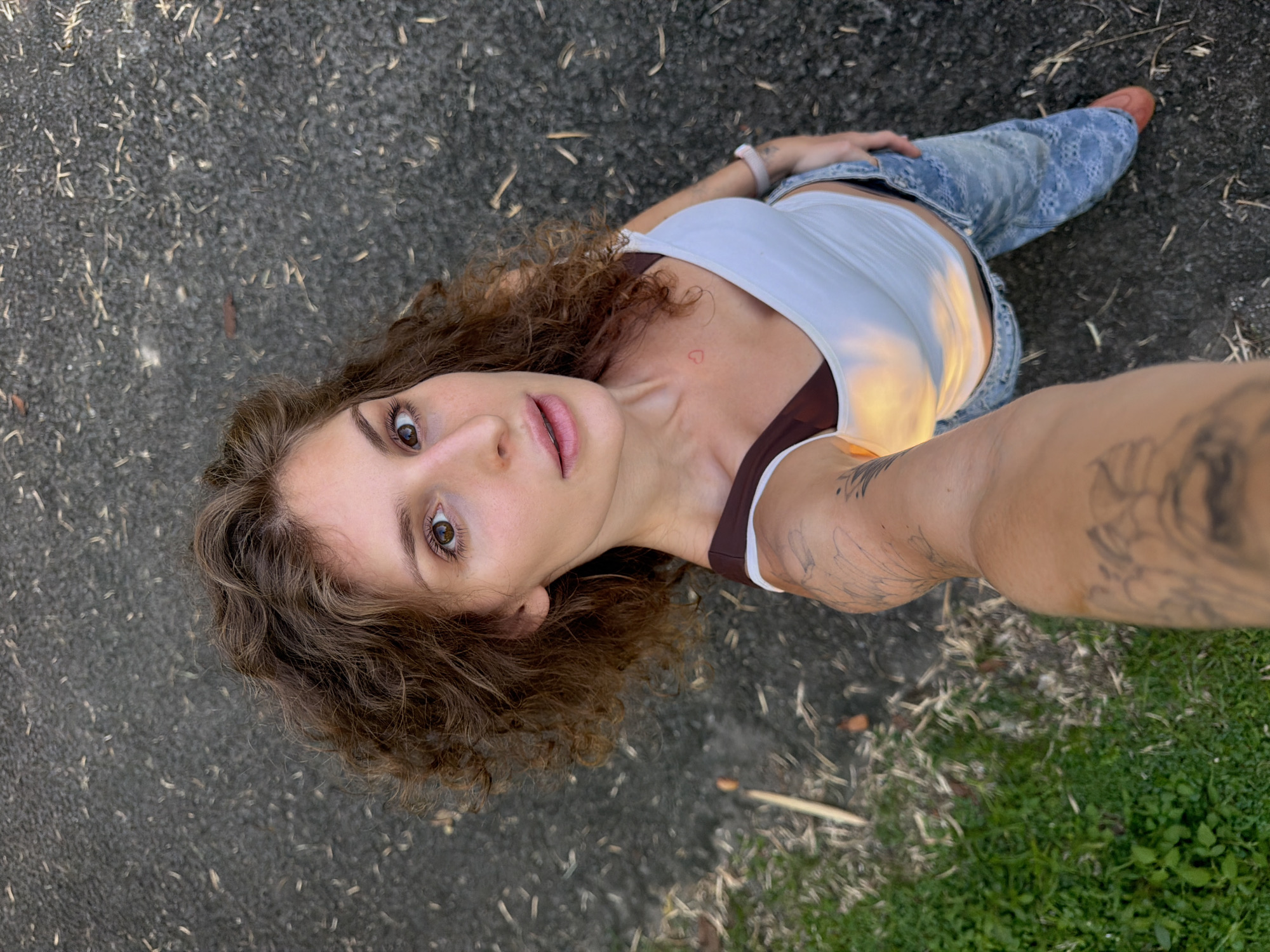 A young woman with curly brown hair is taking a selfie from a low angle, looking up at the camera. She is wearing a white tank top over a brown bra and light blue jeans. A red heart is visible on her chest. Her left arm, displaying tattoos, is extended towards the camera. The ground surface appears to be a mix of asphalt and grass. The location is consistent with outdoor settings found within Kediri, Indonesia.