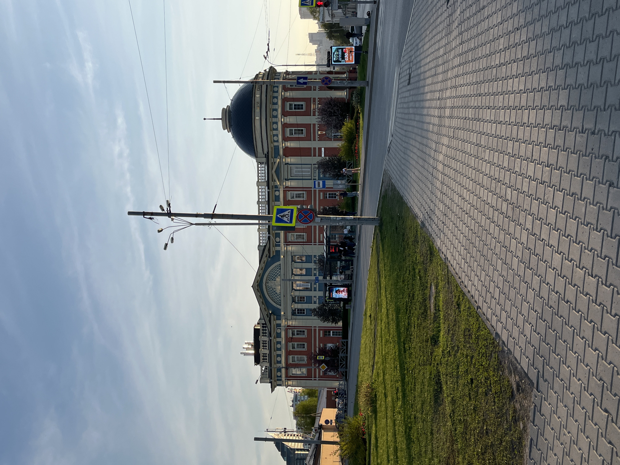 The image captures a street view in Yekaterinburg, Russia, featuring the Sevastianov House as the focal point. The building is a red and blue two-story structure with a large black dome. Utility poles with street lights stand in the foreground. Pedestrian crosswalk signs are visible near the building. A paved sidewalk runs alongside a grassy area. A digital advertisement screen is positioned near the road. The sky is mostly clear with light clouds.