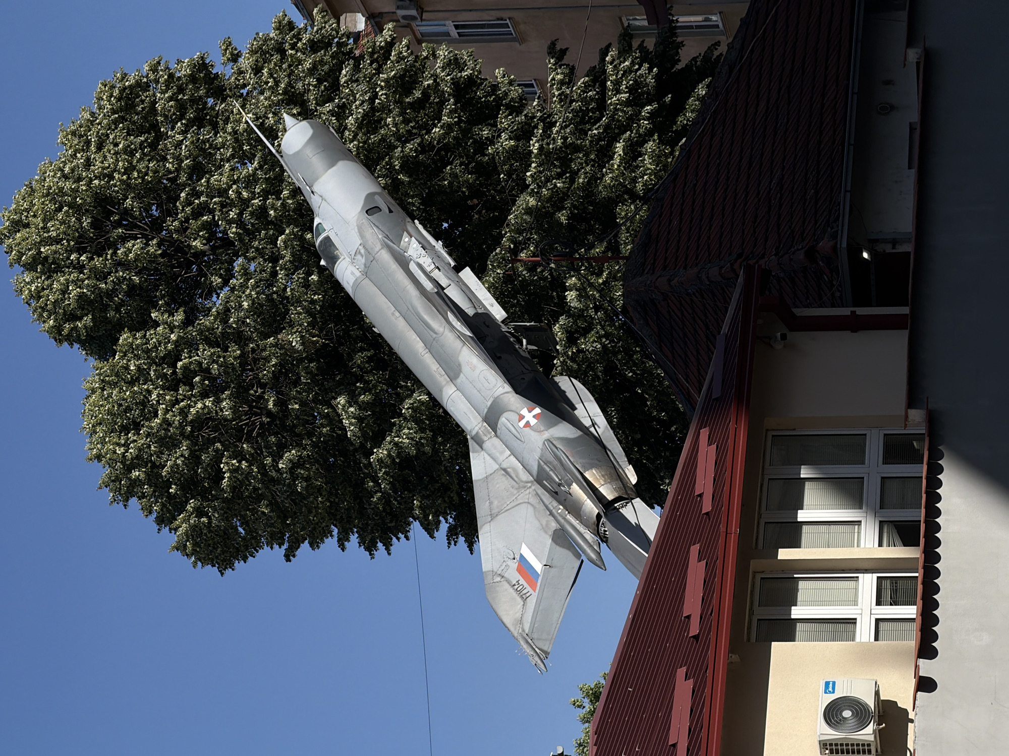 A fighter jet, likely a MiG-21, is mounted vertically as a monument or display in an urban setting. The jet is painted in gray camouflage and marked with a Serbian flag on its tail. It is positioned next to a multi-story building with a red roof and cream-colored walls. The building has windows with white frames. An air conditioning unit is visible on the building's exterior wall. The plane is framed by a large, dense tree with green foliage. The sky is clear and blue. This is believed to be in Belgrade, Serbia.
