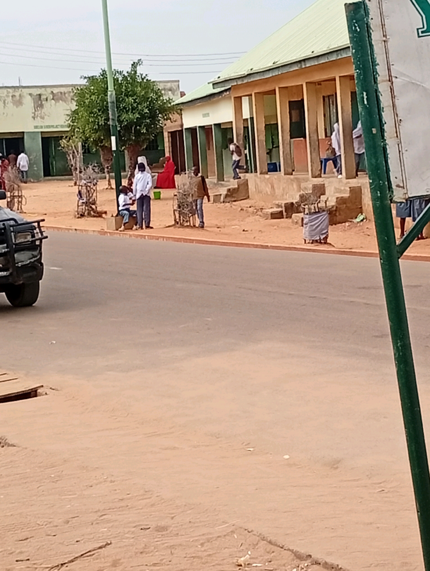 The image depicts a street scene in Gombe, Nigeria, likely a commercial or public area. In the foreground, a paved road dominates the lower half of the frame, with a sandy area and a wooden structure to the left. A dark-colored vehicle is partially visible on the road.
The background features several buildings, suggesting a developed urban environment. One building on the left appears to be a single-story structure with windows and a green door, bordered by a tree. To the right, a building with a light-colored, sloped roof and a covered walkway supported by pillars is prominent. Several people are scattered in front of these buildings.
Individuals are observed standing, sitting, and possibly interacting. Some are dressed in white, others in more colorful attire. There are also objects that appear to be carts or stands with bundles of sticks or materials. A tall green pole, possibly a lamppost or part of a sign, stands vertically on the right side of the image. The overall atmosphere suggests an ordinary day in a busy area, with ongoing activities. The architecture and the presence of a street and buildings are consistent with an urban setting in Nigeria.
