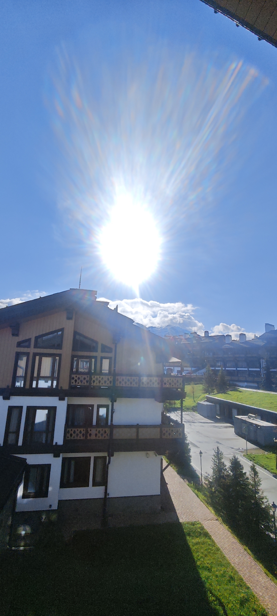 The image shows a multi-story building with a wood and white facade under a bright, sunny sky. The location is Unknown, Russia. The building has multiple balconies with ornate railings. A road runs alongside the building, with street lamps and trees lining the street. Another building is visible in the background, with mountains and clouds in the distance. The sun is very bright, causing lens flare in the image. A brick pathway is alongside the building, and a patch of grass is in the foreground.