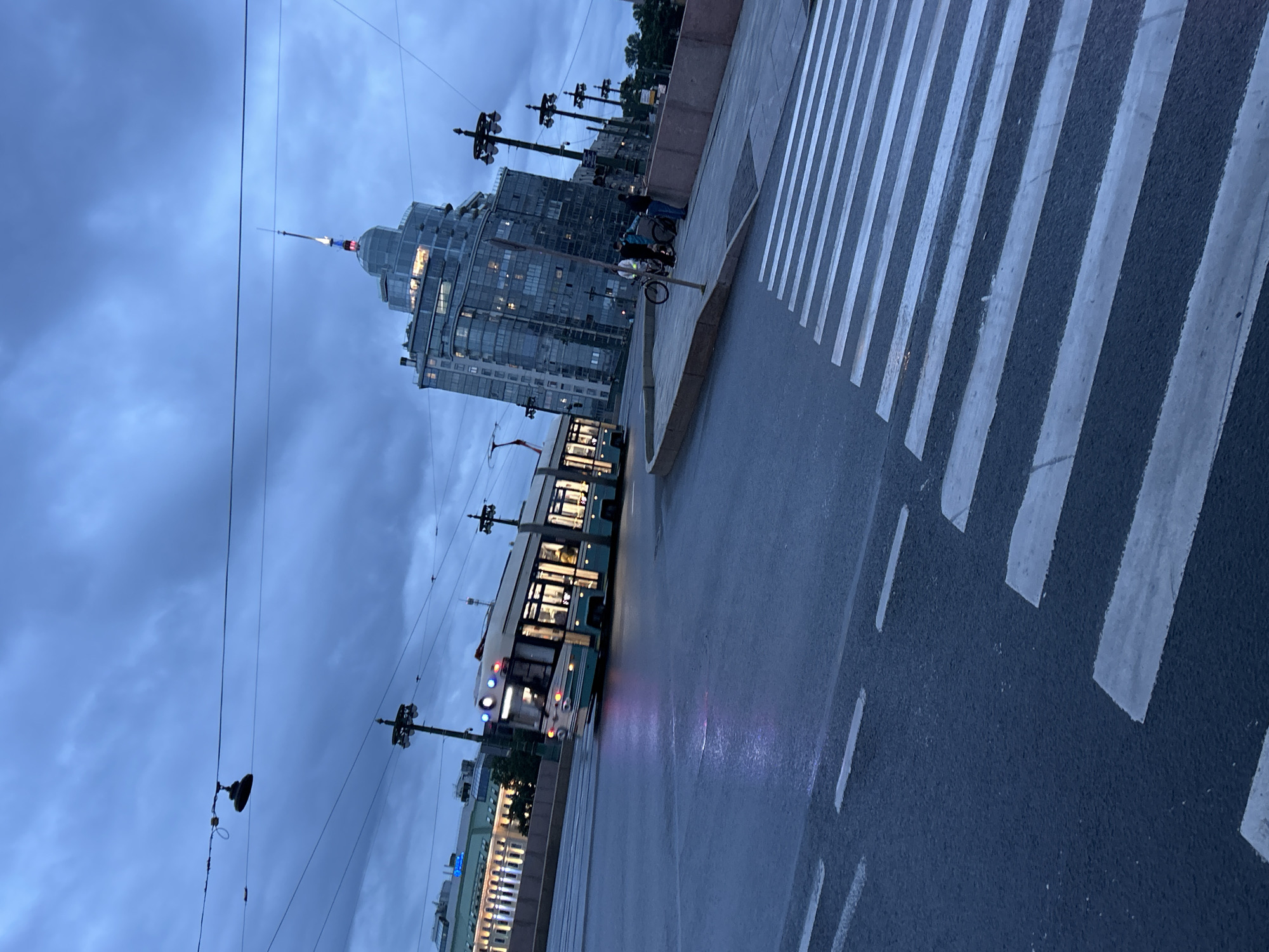 The image depicts a street scene in a city, possibly in the evening or during overcast weather, given the dim lighting and the dramatic sky. A tram, painted in teal and white with illuminated windows, is moving from left to right across the frame, its reflection visible on the wet asphalt. In the background, a distinctive, modern glass building with a pointed spire is prominent. The spire is lit with red and blue lights. Several streetlights with ornate tops are visible along the road and sidewalk. On the right side of the image, a pedestrian crossing is marked with white stripes on the dark asphalt. A few people are standing on the sidewalk near the crossing, one with a bicycle. The overall atmosphere is urban and slightly moody.