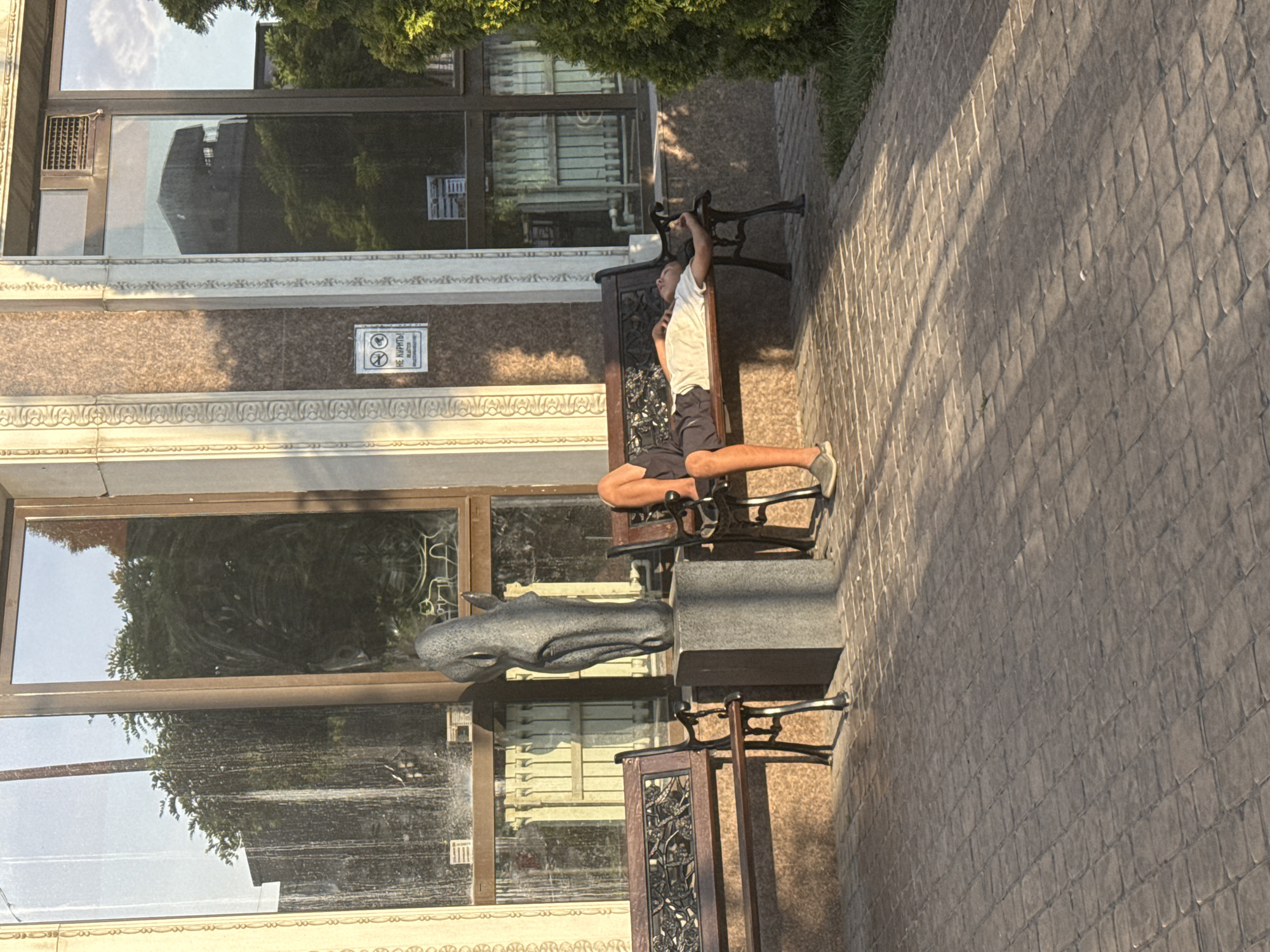 A person is resting on a bench outside a building. The person is lying on their back with their eyes closed, wearing a white t-shirt and dark shorts. Their legs are extended, and their feet are resting on the ground. To the left of the person is a dark metal bench with ornate detailing on the backrest. Next to that bench, and in front of a large window, is a stone sculpture of a horse's head. The building has large windows that reflect the surrounding trees and sky. There is a patterned sidewalk in the foreground. A sign with a "no smoking" symbol and text in Cyrillic is visible on the building's facade. The scene appears to be a public space in Bishkek, Kyrgyzstan.