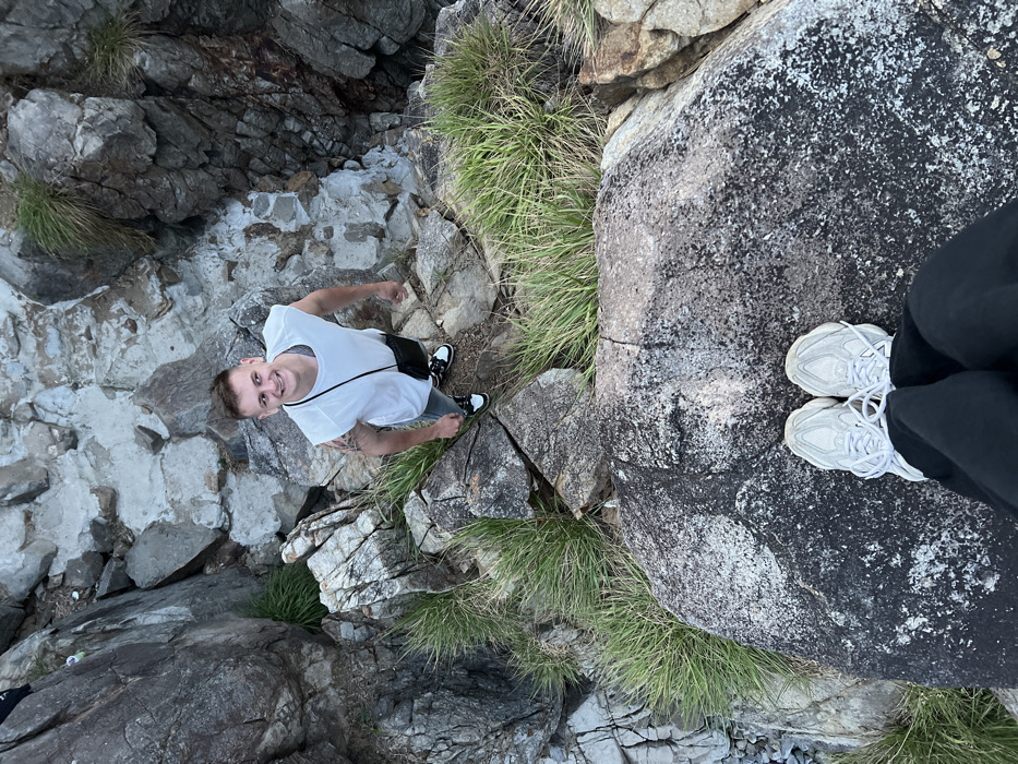 The scene depicts a rocky outdoor environment in Vĩnh Lương, Vietnam, observed from an elevated perspective looking downwards. In the immediate foreground, the lower legs and feet of one individual are visible, clad in black pants and white athletic shoes, standing on a large, dark grey rock surface. Below this position, a male subject stands on a lower, uneven rock formation. He is smiling and looking upwards towards the camera. This individual wears a white sleeveless shirt, light grey pants, and black athletic shoes with white soles. A small black cross-body bag is slung across his chest. Tattoos are visible on his left arm. The surrounding terrain consists of numerous irregular grey and dark grey rock formations, with patches of green grass and weeds growing between them.