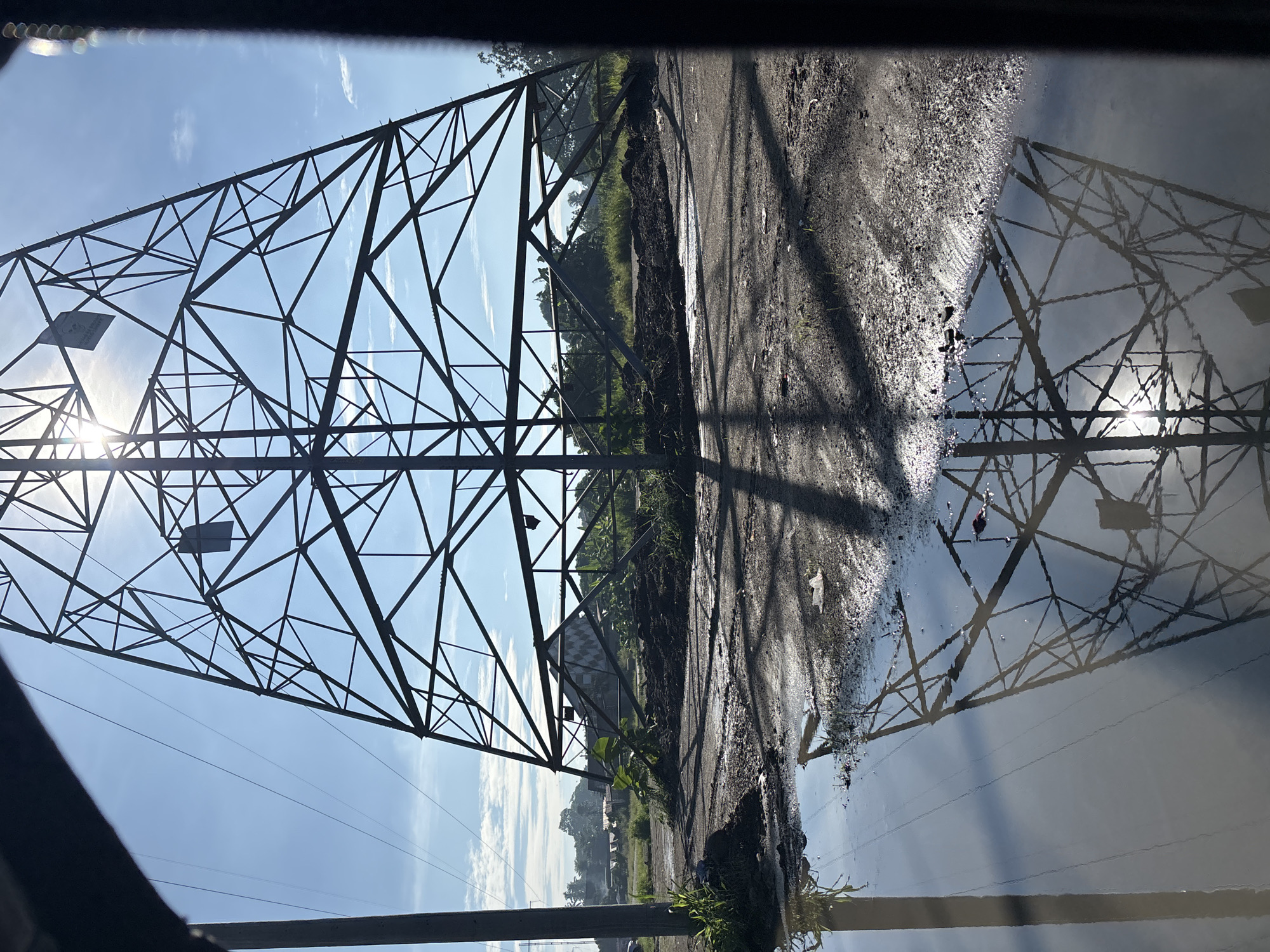 The image shows a partially constructed or damaged power transmission tower located in Tagum City, Philippines. The tower is made of metal lattice and is positioned in an outdoor area with rough terrain, including dirt, mud, and puddles of water. The sky is clear with some scattered clouds, and the sun is visible, casting shadows on the ground. In the background, there are some trees and structures that appear to be residential or agricultural. The scene suggests an ongoing construction project or a site that has been affected by some event. There are no people visible in the image.