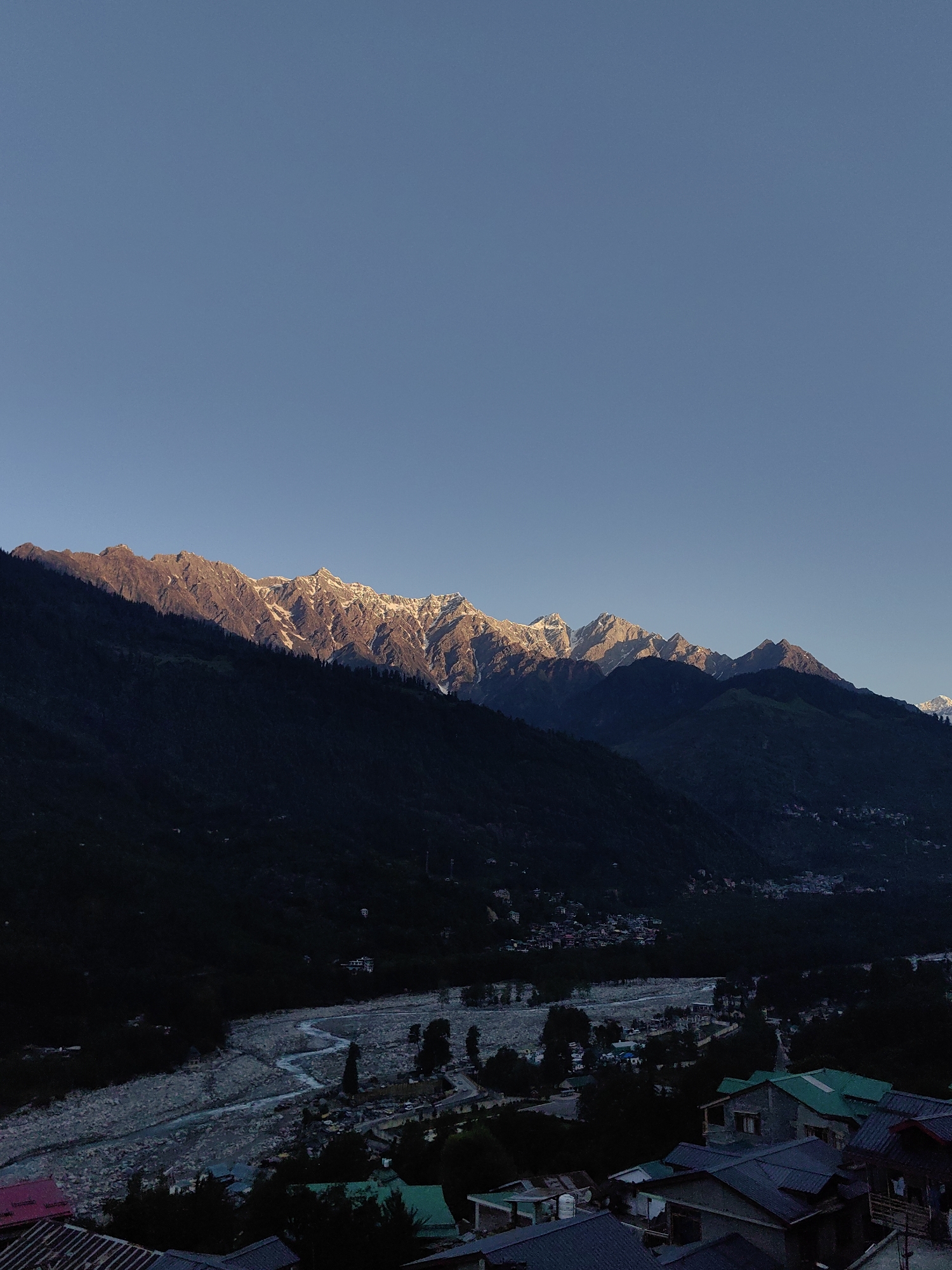 The image depicts a panoramic view of the mountains surrounding Vashisht, India, at what appears to be either dawn or dusk. The sky is a clear, gradient blue, transitioning from a lighter shade at the horizon to a deeper hue overhead. The sun's rays illuminate the snow-capped peaks of the mountain range, highlighting their rugged texture and casting long shadows across their slopes.
In the mid-ground, a lush green mountain slope descends, dotted with scattered trees. A riverbed, appearing wide and stony, winds its way through a valley. Along the river and on the lower slopes, a small settlement is visible, characterized by a cluster of buildings with brightly colored roofs, predominantly green and blue. Some of these structures are closer to the viewer in the foreground, with visible rooftops and architectural details. The overall scene is one of natural beauty, with human habitation nestled within a dramatic mountainous landscape. There are no people or animals clearly visible in the image.