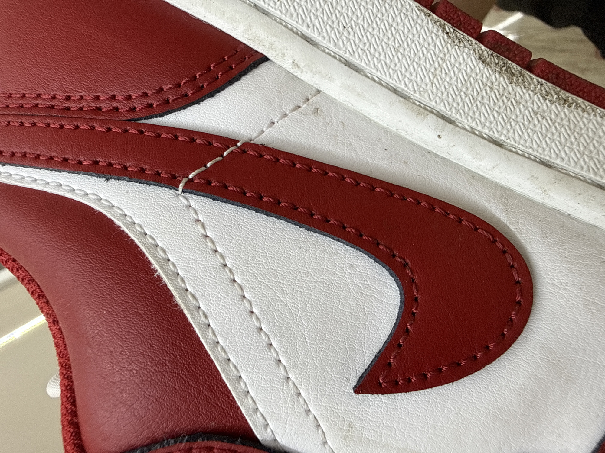 The image is a close-up, high-angle shot of a red and white sneaker, focusing on the side panel and the swoosh logo. The sneaker appears to be made of leather, with white stitching visible along the seams of the red and white panels. The red swoosh logo is prominent, outlined with stitching. The sole of the shoe is white and shows some signs of wear and dirt. The background is out of focus and appears to be a neutral color. There is no indication of the city of Burgas, Bulgaria, or any specific location within it. The image solely depicts a detail of a shoe.