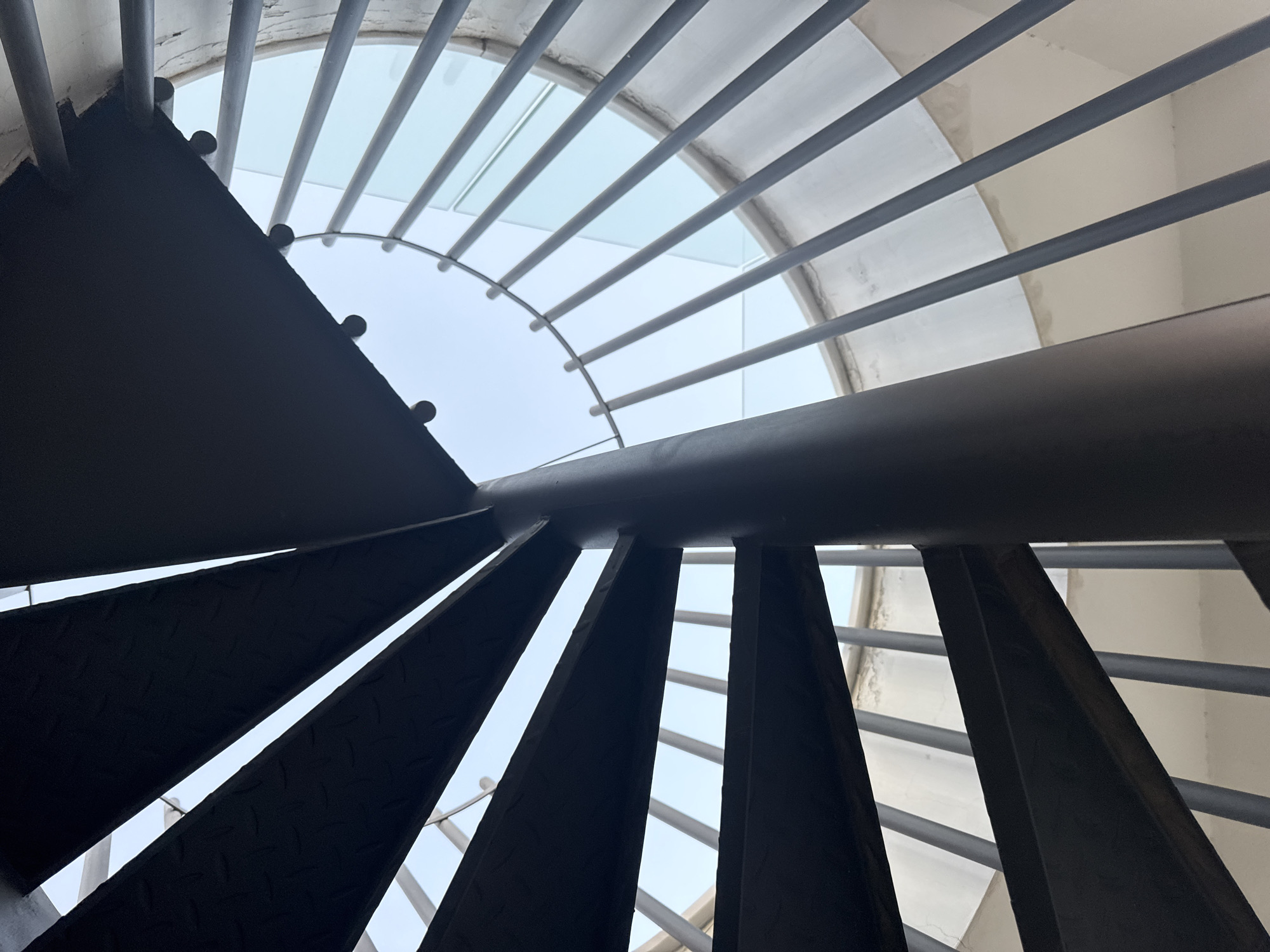 This image shows a close-up, low-angle view of a dark metal spiral staircase leading upwards towards a circular opening in a ceiling, likely an atrium or skylight, in Agios Athanasios Municipality, Cyprus. The staircase has a central dark gray cylindrical pole and the steps are made of dark metal with a textured surface, possibly diamond plate. The sides of the staircase are bordered by a railing consisting of vertical silver metal poles and a curved glass barrier. The background is a bright, pale blue sky, indicating an outdoor or semi-outdoor setting. The surrounding structure appears to be concrete and is painted white. There are no people visible in the image.