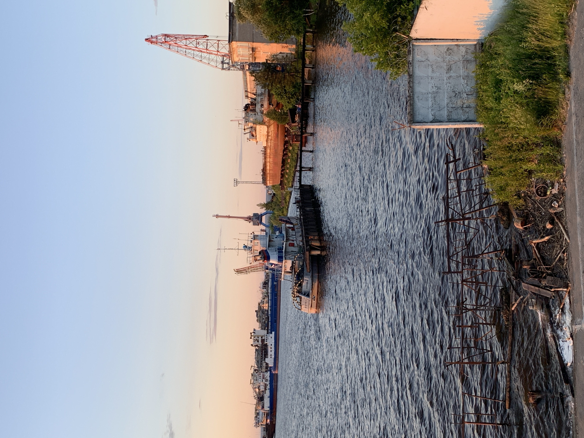 This image depicts a waterfront industrial area in Chaikovsky, Russia, at sunset. The scene is dominated by the Kama River, with several ships and cranes visible along the shore. In the foreground, the dark water is rippled, and a rusty metal structure juts out from the water's edge, with debris and vegetation on the bank. Further out, a tugboat is moored to a wooden pier. In the mid-ground, several large cargo ships are docked, their hulls showing signs of age. To the right, a large red crane stands tall next to a brick building, bathed in the warm glow of the setting sun. Trees and other vegetation are present along the shoreline, adding a touch of nature to the industrial landscape. The sky is clear and transitions from a pale blue at the top to a warm orange and pink hue near the horizon, indicating the time of day. No people are visible in the frame.