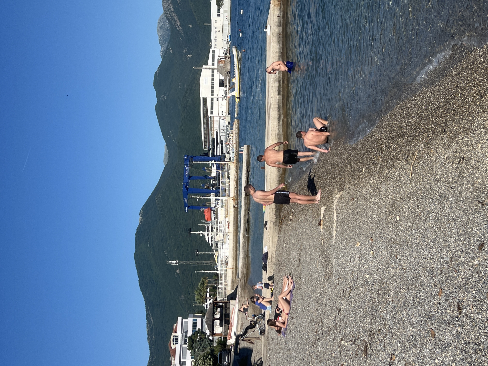 The image captures a scene on a sunny day in Bijela, Montenegro. A small, pebbled beach is populated with people. Several individuals are in the water, some standing near a concrete pier-like structure that extends into the water, while one is swimming further out. On the beach, a group of people are sunbathing on towels. A man stands on the beach close to the water. In the background, there's a harbor with cranes, boats, and buildings. Mountains rise behind the harbor, with a clear blue sky above. A few residences sit near the beach.