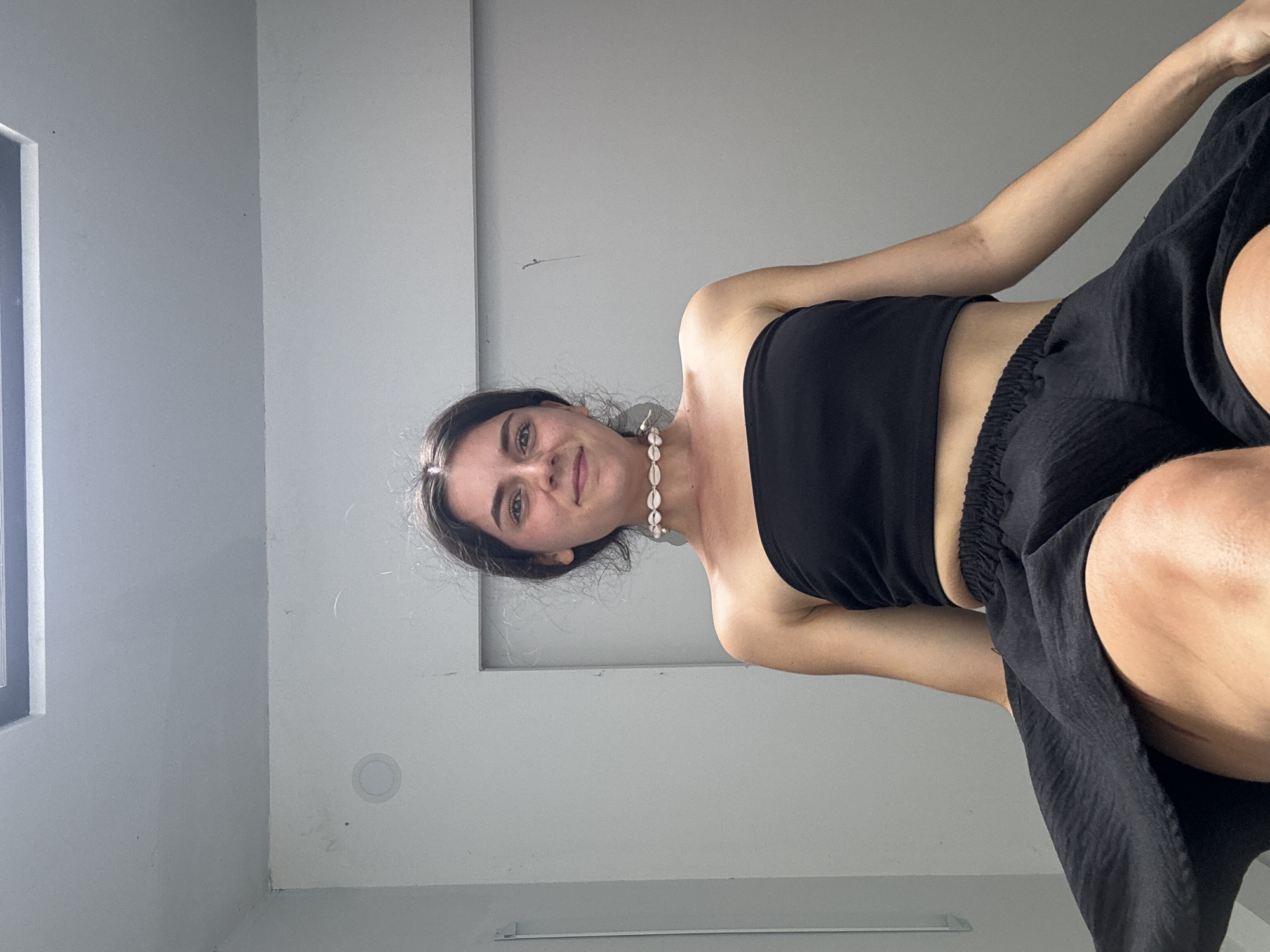 A fair-skinned woman wearing a black tube top and black shorts is looking at the camera. She is also wearing a necklace made of shells. The location is an interior space in Mũi Né, Vietnam. The walls are gray.