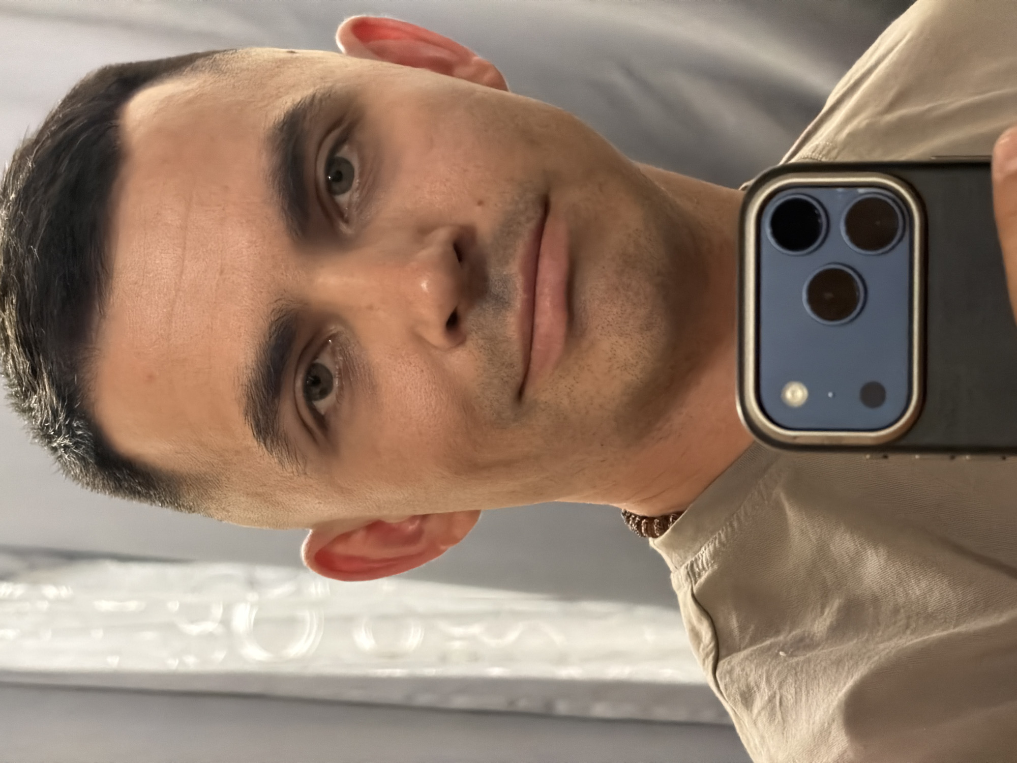 A close-up, portrait-style photograph shows a man holding a smartphone in front of his face as if taking a selfie. The phone obscures part of his face on the right side. The man has short, dark hair, light skin, and light-colored eyes. He is wearing a light-colored shirt. A partial view of a mirrored surface with circular patterns is visible in the lower left corner of the frame. There is no identifiable evidence to indicate this image was taken in the city of Izhevsk, Russia.