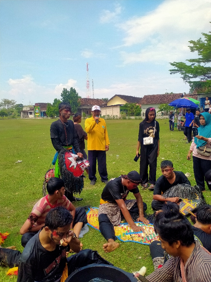 The image depicts an outdoor scene in Jetak, Indonesia, on a grassy field under a partly cloudy blue sky. In the background, several single-story buildings with light-colored walls and red-tiled roofs are visible, along with scattered trees and a prominent red and white cellular tower. To the far right, a blue and white canopy or umbrella is present.
In the midground, a man in a yellow collared shirt, dark trousers, and a white baseball cap stands, holding a microphone to his mouth as if addressing a group. To his left, another man stands, wearing a dark shirt and dark trousers, with red facial markings. He holds a large, elaborate mask or prop featuring a red face, a white five-pointed star eye, and dark shaggy material. To the right of the man in yellow, a woman stands, clad in a black long-sleeved shirt, black trousers, and a white crossbody bag, holding a dark smartphone. A woman in a light blue hijab is partially visible on the far right.
In the foreground and mid-foreground, several individuals, primarily men, are seated on the grass. Many exhibit red facial markings, and their clothing, mostly dark shirts and trousers or sarong-like garments, appears wet. One man on the far left wears a red and white striped shirt. In the center, a man is lying partially on a colorful, woven mat while interacting with it. Another man next to him, also appearing wet and with red markings near his eye, holds a dark, shaggy prop or mask. A man in the immediate foreground, with red facial markings, reaches into a dark, large container or object. The scene suggests a break or preparation period during a traditional performance or cultural event.