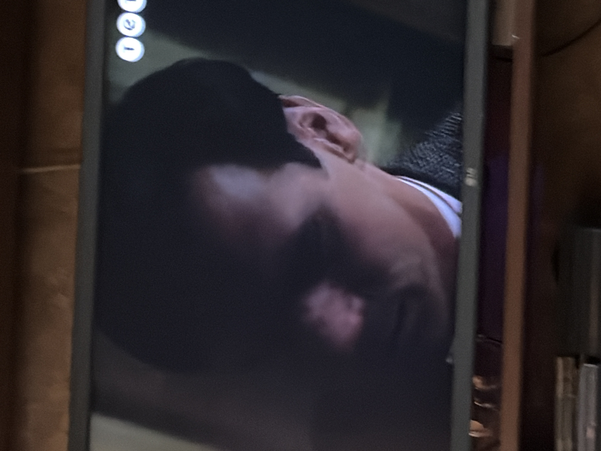 The image shows a close-up of a television screen displaying a man's face. The man has dark hair and is looking off to the side. His face is partially obscured by shadow, but his ear and the side of his head are visible. He is wearing a dark jacket over a white shirt. The television screen is mounted on a shelf, and the top of the shelf and a portion of the wall are visible in the background. There are some circular icons in the upper right corner of the television screen. The image is grainy and out of focus, suggesting it was taken with a camera or phone from a distance. There is no identifiable information in the image that would suggest a location in Osolynka, Ukraine.