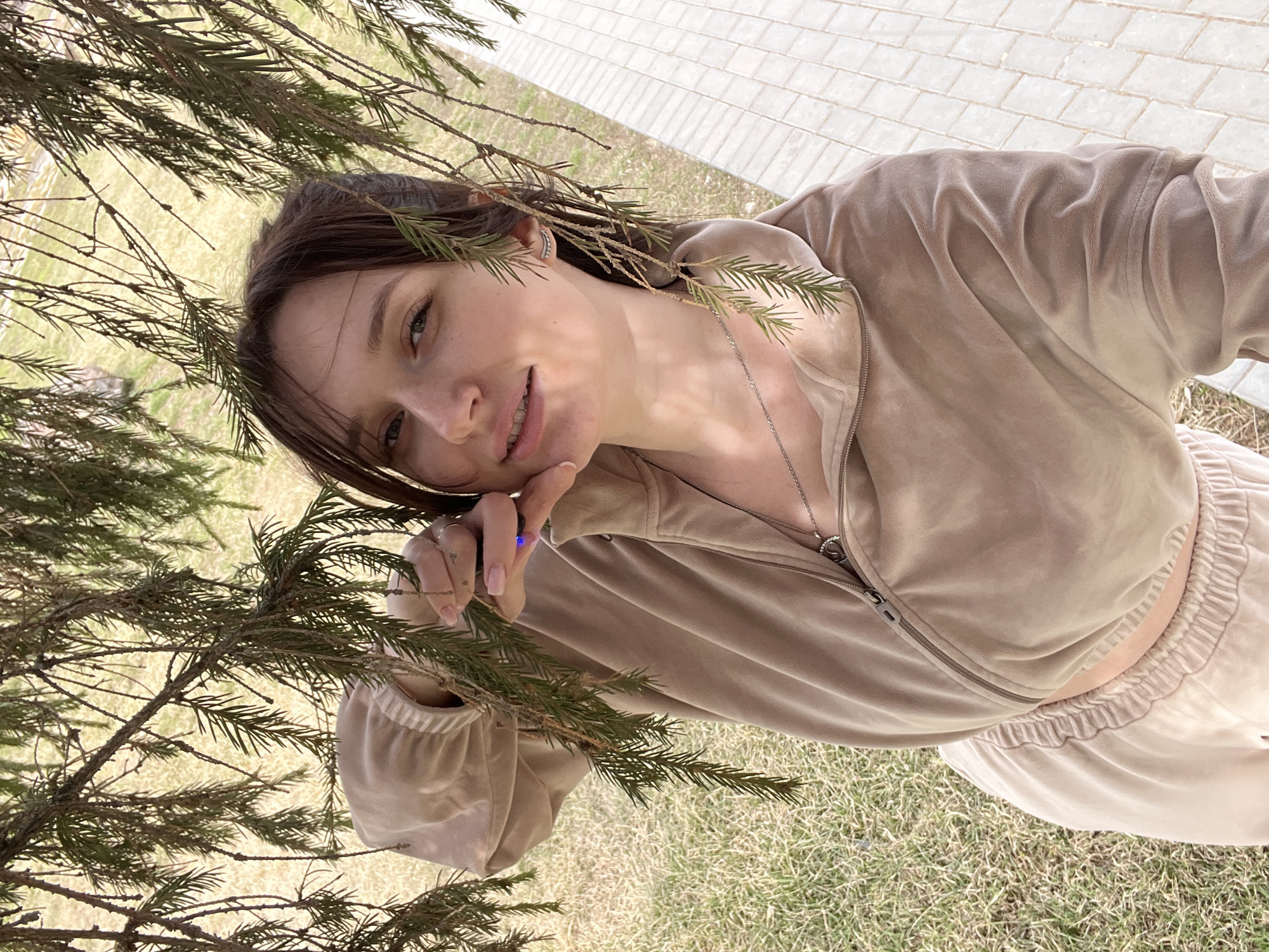 A young woman in Kargapolye, Russia, poses outdoors under the branches of a coniferous tree. She wears a beige velour tracksuit with a zippered jacket and matching pants. Her dark hair is styled with some branches tucked behind her ear. A silver necklace is visible around her neck. She is holding a tree branch with her hand, which has manicured nails. The background shows a paved walkway and grassy areas.