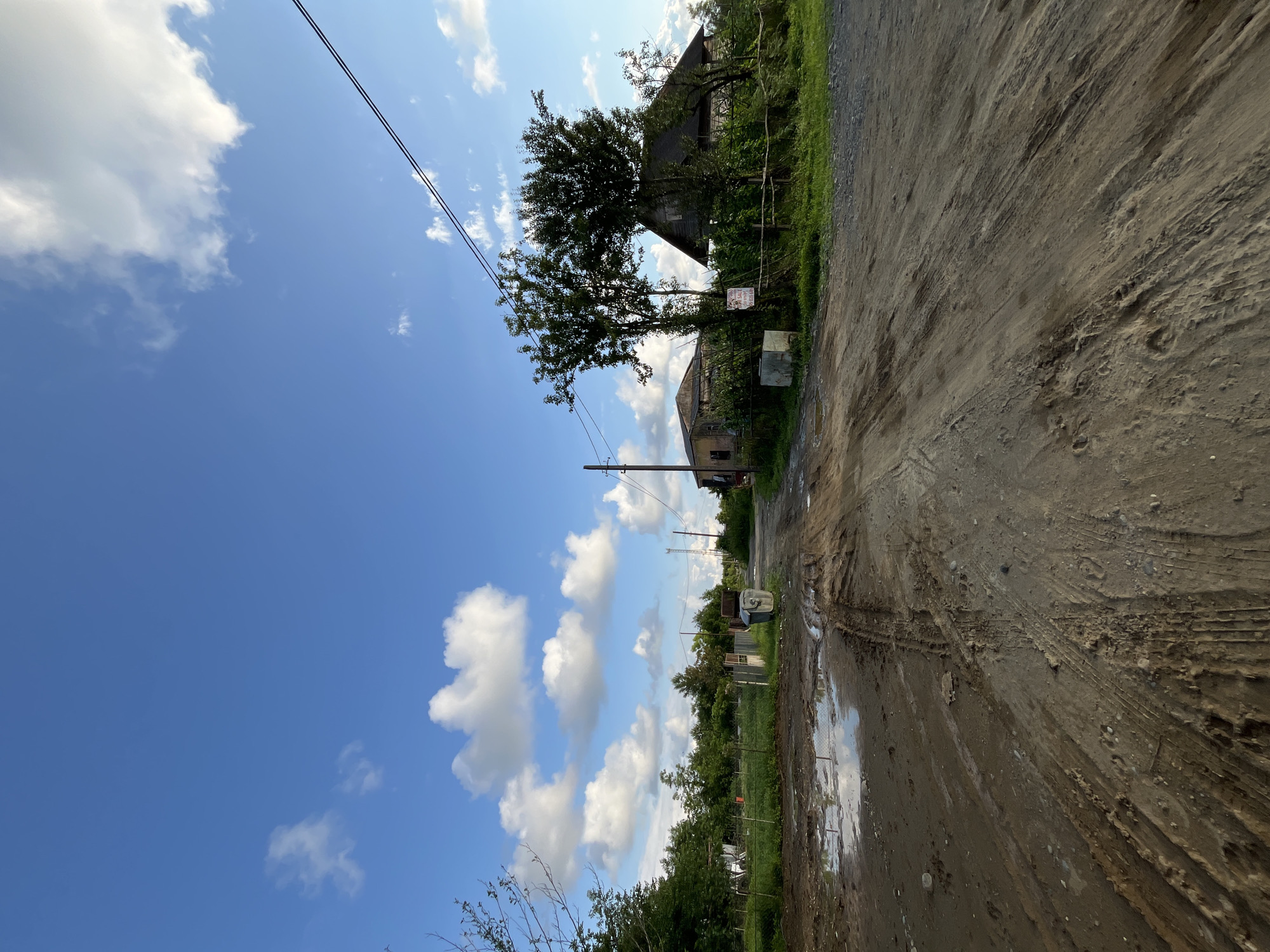 The image depicts a rural dirt road in Ureki, Georgia. The road is muddy and appears to have been recently rained on, with puddles of water reflecting the sky. Tire tracks are visible in the mud, indicating vehicle passage. On the left side of the road, there is a grassy field enclosed by a fence, with some small structures and trees. A large trash bin is also present near the fence.
Further down the road, on the right side, two houses are visible. The house in the foreground on the right is a wooden structure with a sloping roof, partially obscured by trees and vegetation. A "For Sale" sign is affixed to a post near this house. Behind it, another building with a lighter colored facade is visible. Power lines stretch across the top of the frame, supported by a utility pole. The sky is blue with scattered white clouds, suggesting fair weather. The overall scene conveys a sense of a quiet, possibly underdeveloped, residential area in a rural setting.