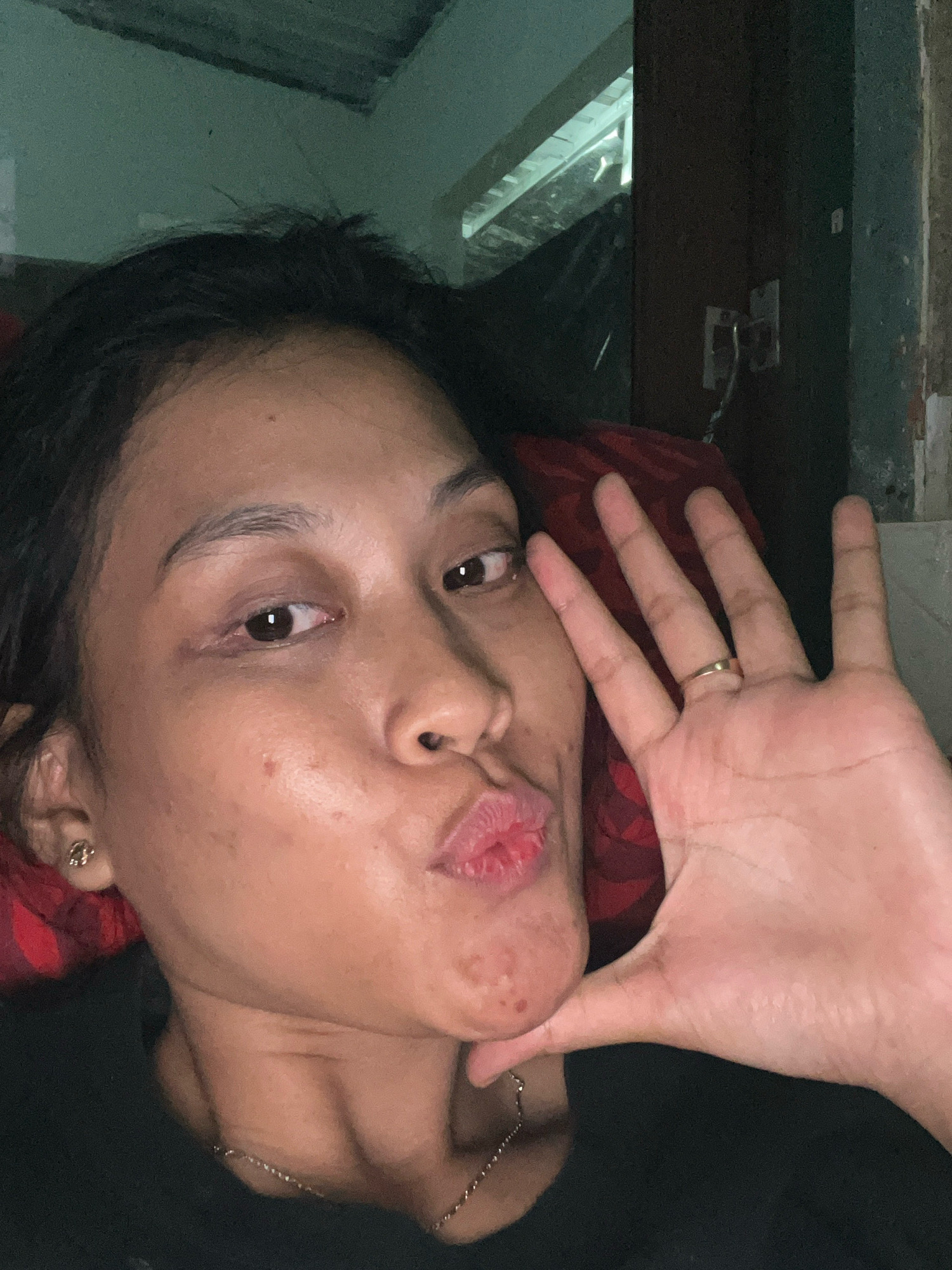 The image is a close-up, indoor, selfie-style photograph of a person in Kecamatan Pati, Indonesia. The subject, a woman with dark hair and brown eyes, is looking directly at the camera with her lips pursed. She is wearing a black top and a thin silver necklace. Her right hand is raised, with fingers spread, and she is wearing a gold ring on her ring finger. The background is dark and somewhat blurry, with glimpses of a ceiling, a doorway, and what appears to be red fabric. The lighting is dim, casting shadows on the subject's face.