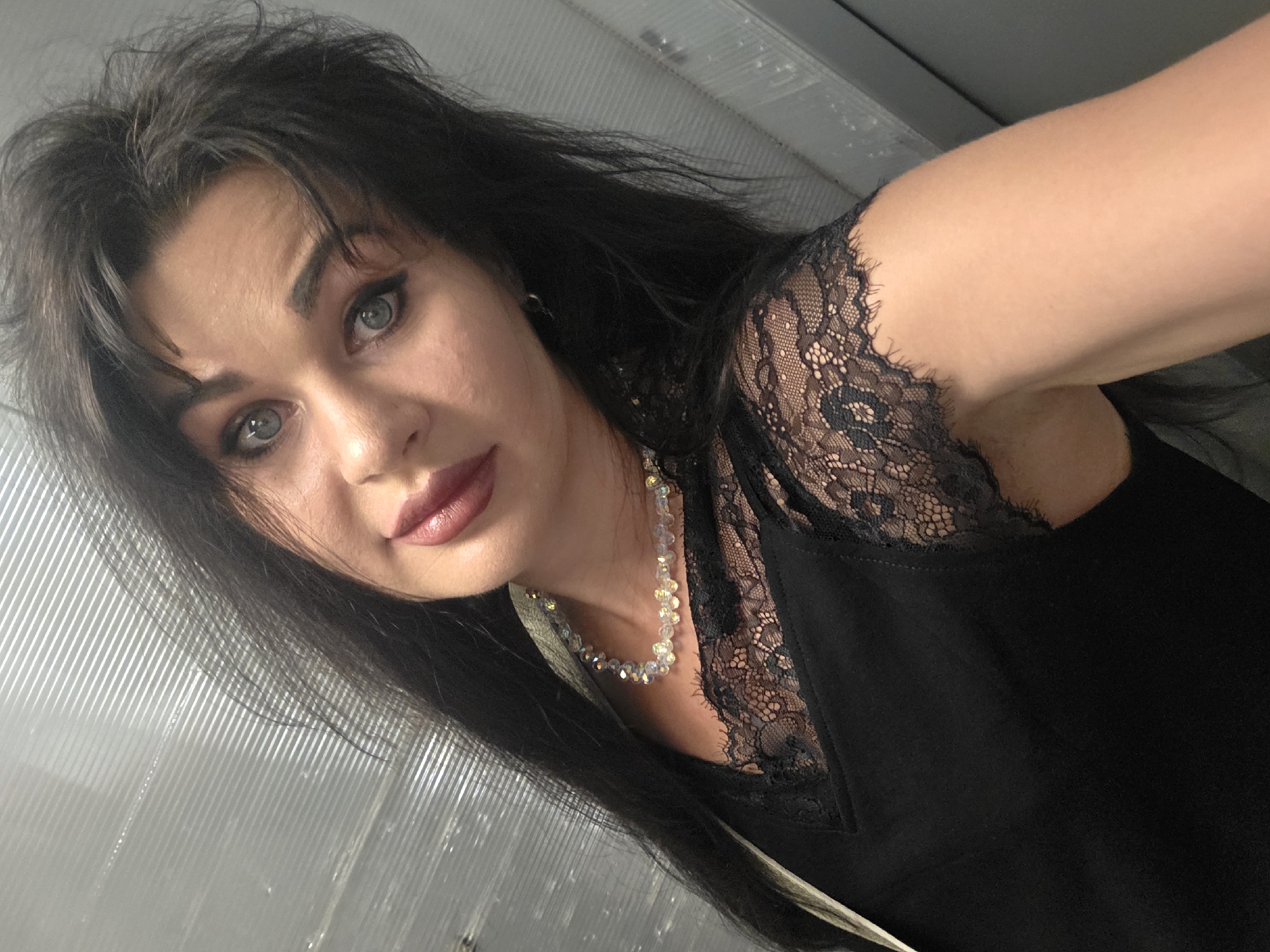 A woman with long dark hair and striking blue eyes takes a selfie in a somewhat cramped space. She is wearing a black top with delicate black lace trim on the neckline and sleeves, and a beaded necklace. Her makeup is noticeable, with dark eyeliner and lipstick. The background appears to be a textured, metallic wall, possibly in a public restroom or changing area. The lighting is somewhat harsh, creating reflections on the metallic surface. There is no clear indication of the specific location within Sochi, Russia, but the environment suggests an indoor, utilitarian setting.