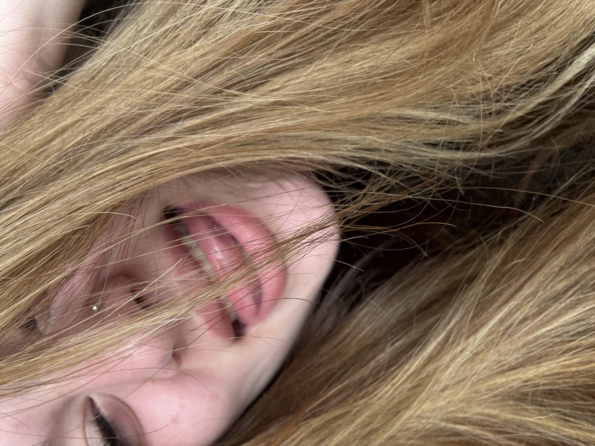 The image shows a close-up of a person's face obscured by long, light brown or blonde hair. Part of their face is visible, including an eye, cheek, ear, and mouth with teeth slightly parted. The overall scene is intimate and focuses primarily on the texture of the hair and the exposed portion of the face. There are no contextual details that suggest the location is Troyan, Bulgaria, or any other specific place.