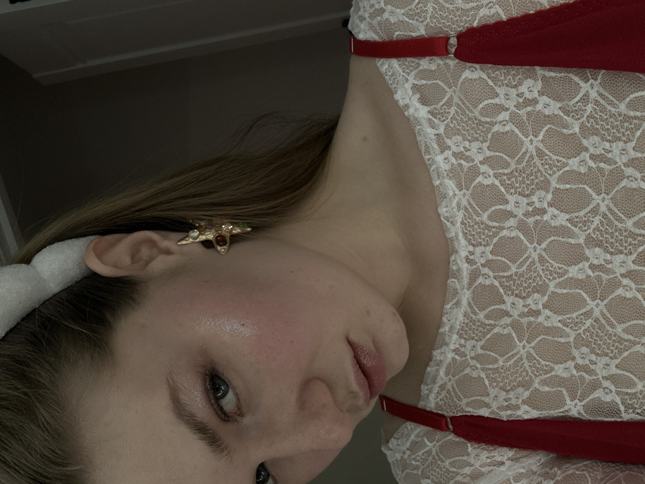 A woman's face, angled slightly, features a gold, multi-colored gemstone earring shaped like a star. A white headband sits atop her blonde hair. Her skin is fair, with a hint of pink in her cheeks. She wears a white lace garment with red straps. The background is muted and dark.