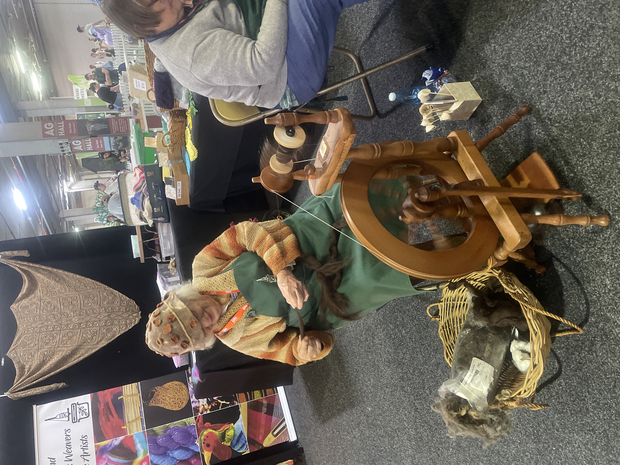 The image shows a woman in her late 70s or 80s with white hair, wearing a knitted hat decorated with floral appliqués, a yellow and orange striped cardigan, and a green apron. She is seated and actively engaged in spinning yarn using a wooden spinning wheel. Her hands are occupied with dark brown wool. The spinning wheel is made of polished wood and has a large wheel and a spindle mechanism. Beside her, to the left, is a woven basket containing bags of wool and some small white cotton. In the background, a display banner with images of crafts and the text "and Weavers Artists" is visible. To the right, another person, partially visible, is seated in a chair, wearing a grey top and blue pants. The environment appears to be an indoor exhibition hall, possibly a craft fair or agricultural show, with a dark carpeted floor and the presence of other stalls and attendees in the distance. The overall scene depicts a demonstration of traditional textile craft.