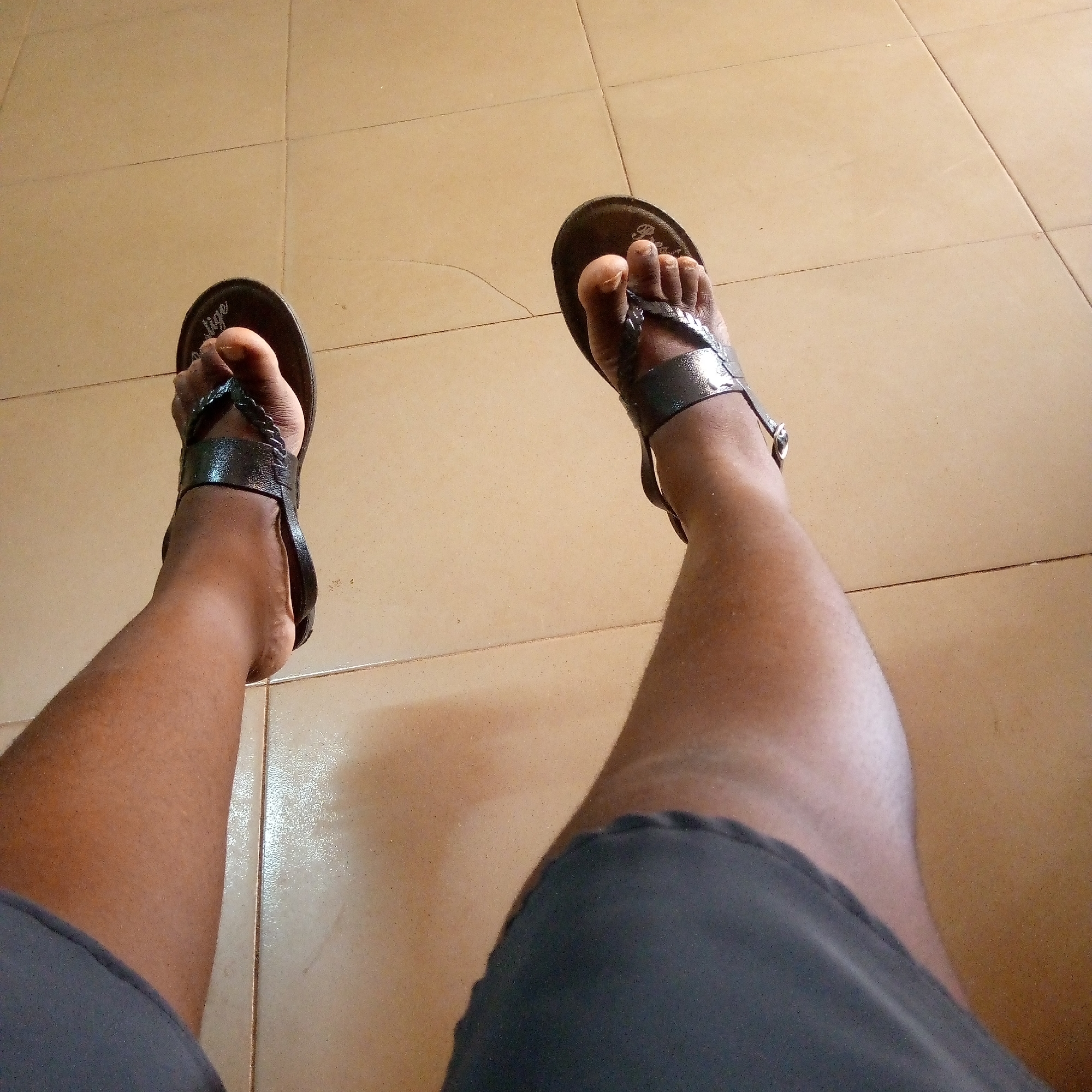 A first-person perspective shot shows a person's legs and feet wearing black sandals. The person is wearing gray shorts. The background consists of beige-colored tiled flooring. The location is Unknown, Unknown.