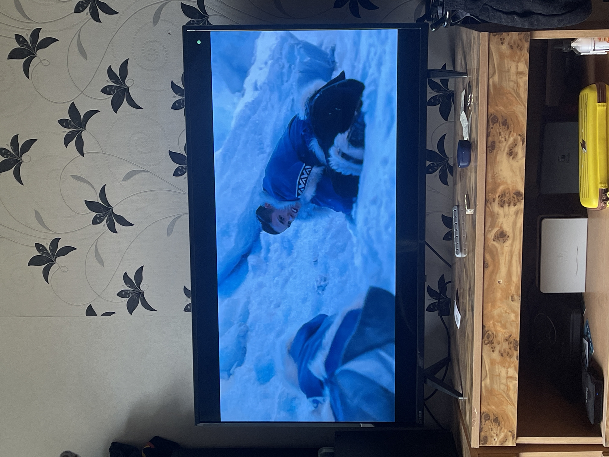A television screen displays a scene from a movie set in an icy environment. A young man with dark hair is depicted lying in snow or ice, wearing a blue garment with white fur trim and geometric patterns around the neckline. He appears to be distressed or in a struggle. The background of the screen shows more of the snowy landscape. The television is placed on a wooden cabinet. On the cabinet are various objects, including remote controls, a blue circular object, and some papers. Below the cabinet, several electronic devices are visible, along with a yellow plastic case. The wall behind the television is adorned with floral wallpaper featuring dark grey flowers and vines. The overall scene suggests an indoor setting in a residential space. The location is not specified within the image.