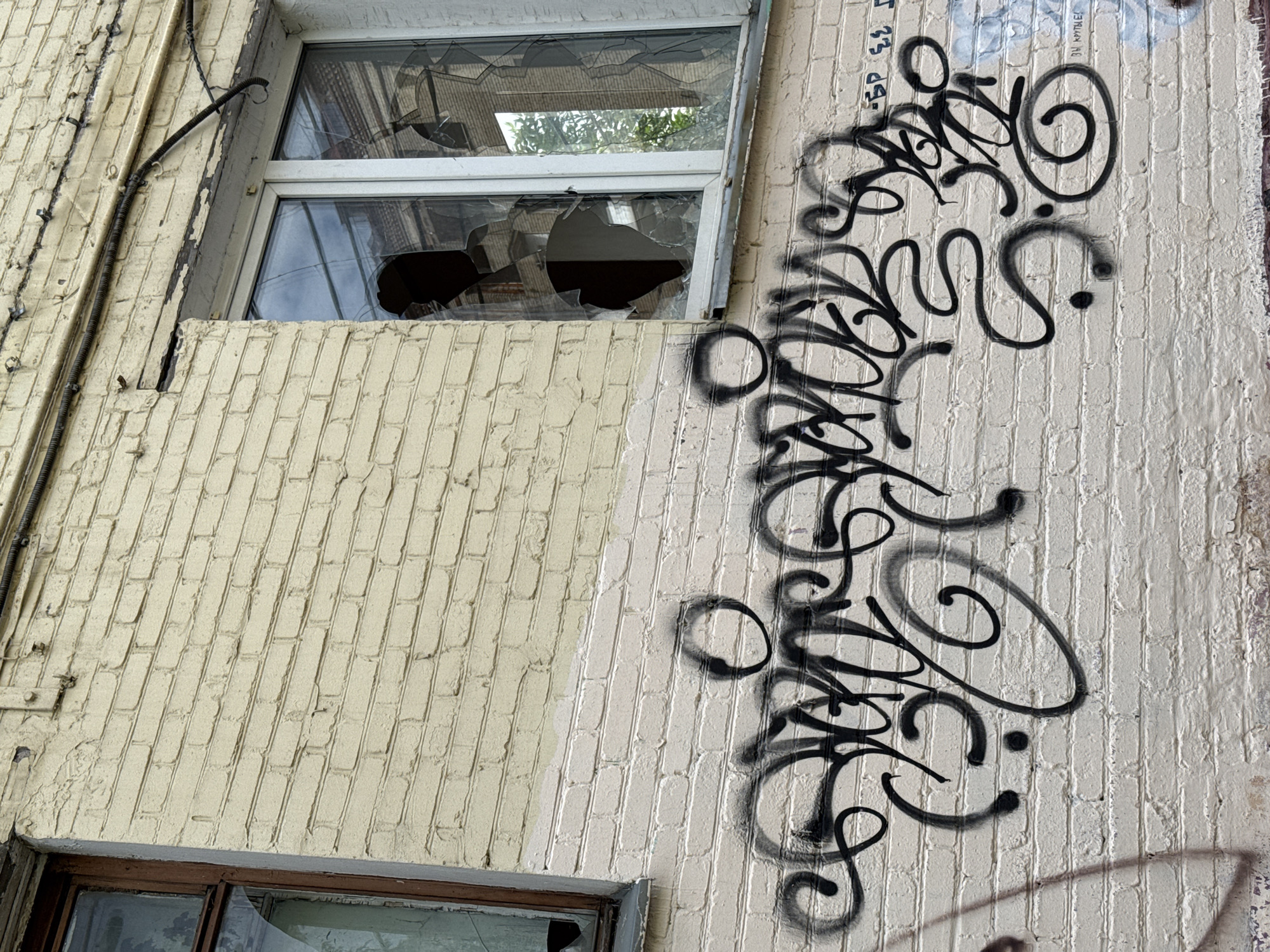 The image shows a section of a brick building wall in Moscow, Russia, with graffiti art. The wall is painted a light yellow on the upper portion and off-white on the lower portion. There are two windows visible; the left window has a brown wooden frame and cracked glass, while the right window has a white frame and is severely broken, with large shards of glass missing. Black graffiti, in a stylized script, covers the lower, off-white section of the wall. Electrical conduits run along the top of the wall above the windows.