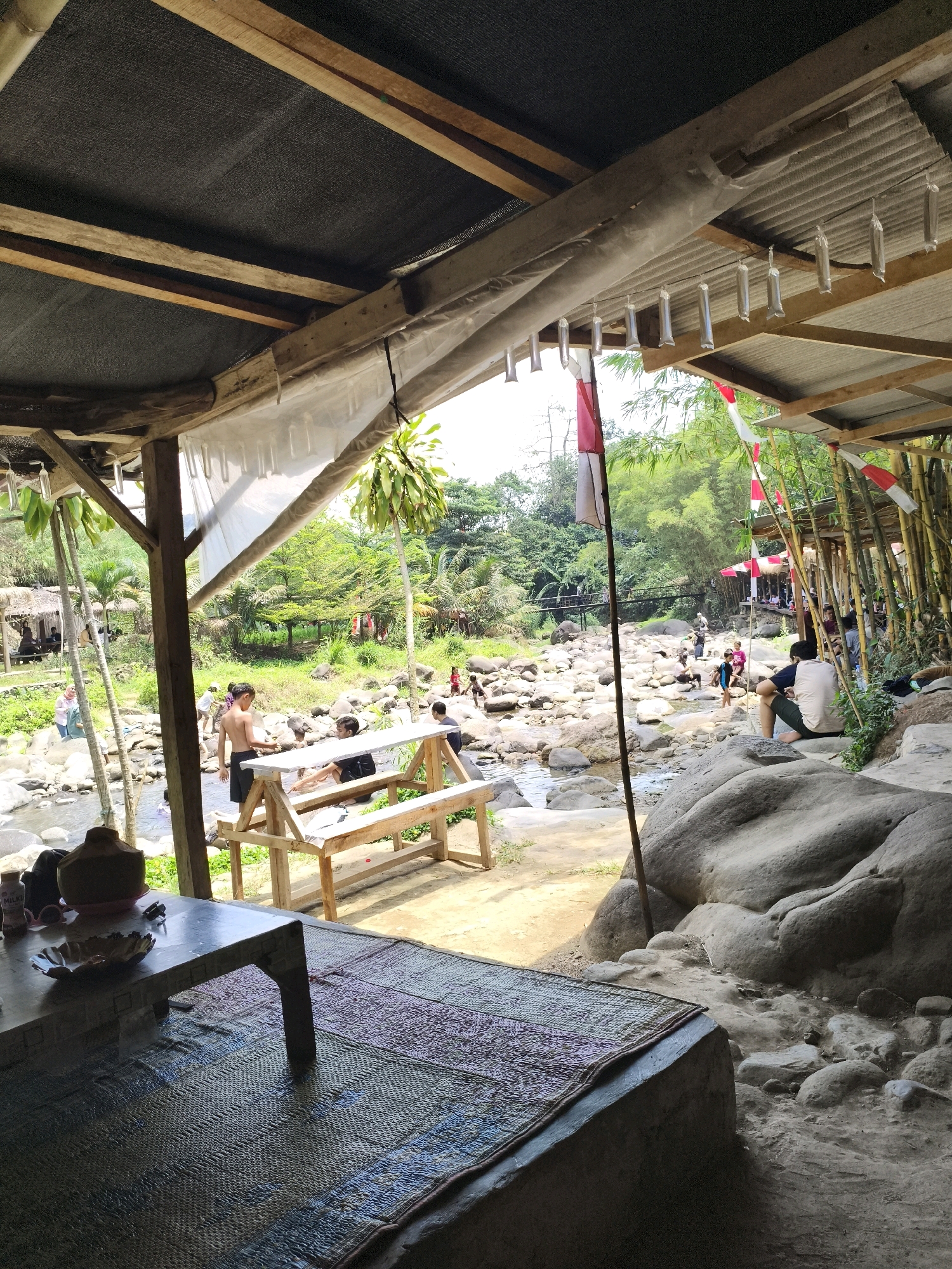 In Babakan Madang, Indonesia, an outdoor dining and relaxation area is situated adjacent to a rocky riverbed. A wooden structure with a corrugated metal roof, covered in part by a black mesh material, provides shade. Hanging from the ceiling are clear plastic bags filled with water. Red and white Indonesian flags are displayed.
In the foreground, a low table with a patterned mat is set on the ground. A coconut and other items are on the table. To the right, large boulders form part of the landscape.
Beyond the immediate seating area, people are seen in and around the river. Some are sitting on rocks, while others are wading or playing in the shallow water. A wooden bench and picnic table are positioned near the riverbank. Lush green trees and vegetation surround the river and extend into the background. A small bridge can be seen in the distance.
Several individuals are visible, including a young man standing shirtless in the river, and a man sitting on a rock. Other figures are scattered throughout the scene, engaged in various activities. The overall impression is of a casual, natural recreational spot.