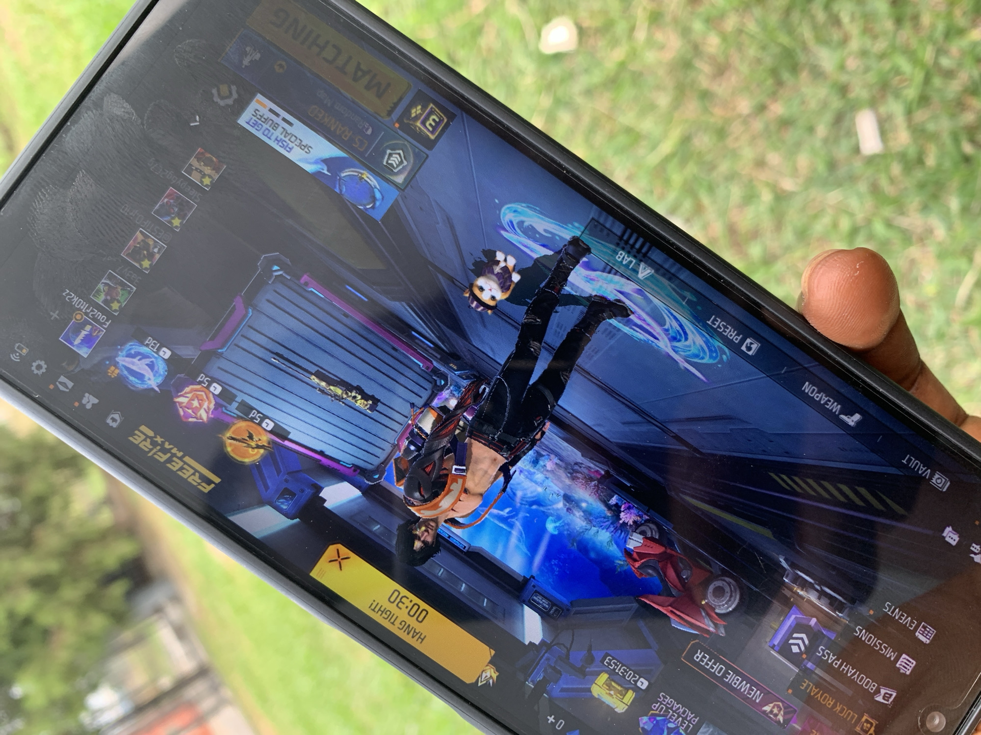 A close-up shot shows a person holding a smartphone, angled slightly. The screen displays the Free Fire Max mobile game interface, featuring a character and in-game menus. The background behind the phone is blurred green foliage, suggesting an outdoor location. The person's thumb is visible on the edge of the phone. No buildings or identifying landmarks are visible to confirm the location. The image does not provide enough information to confirm the city of Sangotedo, Nigeria.