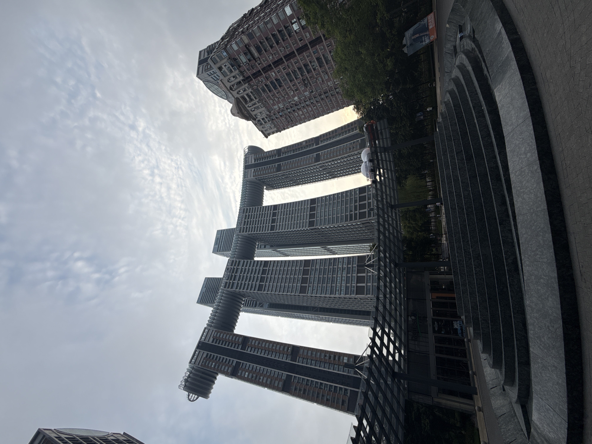 A low-angle, wide shot captures a dynamic urban scene in Yuzhong District, China, dominated by striking modern architecture under a cloudy sky. In the foreground, a series of tiered, dark gray stone steps curve towards a shaded entrance area with a glass-paneled roof supported by dark metal beams. The steps appear to be part of a water feature or amphitheater. Behind the entrance, a transportation system with a red and white vehicle is visible on an elevated track.
The main architectural marvel of the image is a cluster of towering skyscrapers. Three slender, tall buildings stand in the center, connected at their upper levels by massive horizontal structures that create a distinct, almost arch-like form. These central buildings are clad in glass and metal panels, reflecting the overcast sky. To the left and slightly further back, another skyscraper leans dramatically outwards, also connected to a central structure by a similar horizontal element. This building features a mix of dark glass and textured, lighter-colored panels. To the right of the central cluster, a taller, brick-red building with a more traditional, ornate facade rises, featuring numerous windows and decorative architectural details. Lush green trees are visible in the mid-ground between the foreground and the buildings, and a large advertisement poster is propped up on the right side of the image. The sky is filled with wispy white clouds, suggesting a partly cloudy day. There are no visible individuals in the foreground or mid-ground.