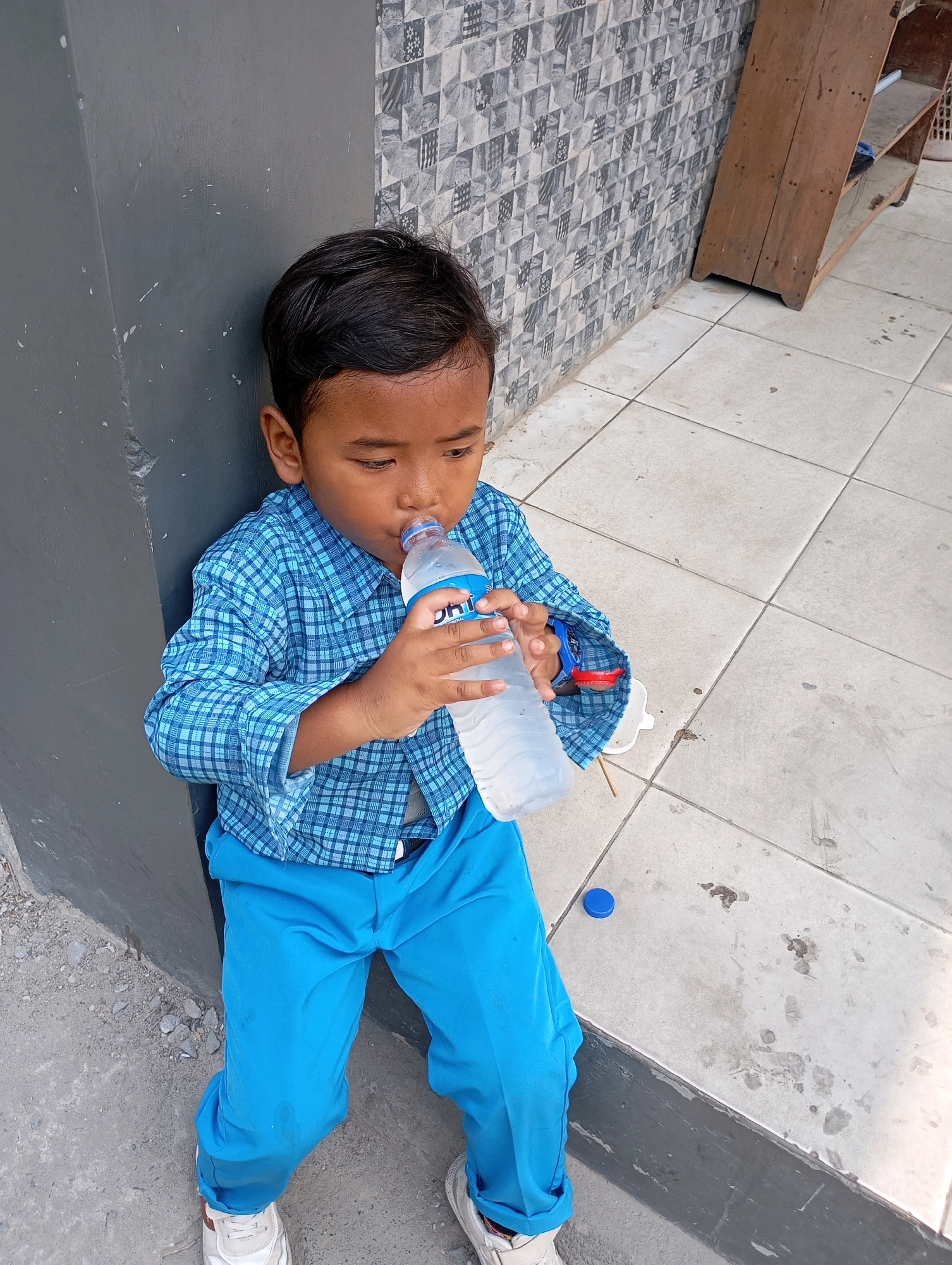 A young boy, appearing to be of Asian descent, sits leaning against a gray wall. He is wearing a blue plaid shirt and bright blue pants, with white sneakers on his feet. The boy is holding a clear plastic bottle of water and drinking from it. The background shows a tiled wall with a geometric pattern, a wooden shelf unit, and a tiled floor. A blue bottle cap lies on the floor near the boy. The scene appears to be outdoors, possibly in a public area within the city of Babelan, Indonesia, given the context provided.