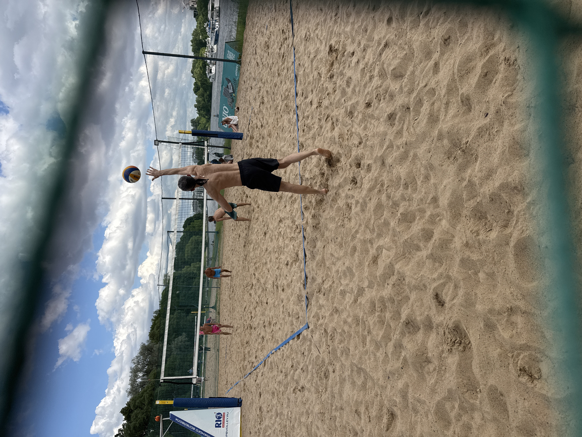 The image depicts a beach volleyball game in Saint Petersburg, Russia, likely at a venue such as "Beach Volleyball Club Rio" or a similar facility along the Neva River embankment. The court is made of sand, with a net dividing it. In the foreground, a shirtless male player in black shorts is captured mid-action, tossing a volleyball into the air with his left hand, preparing to serve or spike. His back is to the camera, and he has a ponytail. Further back on the court, another male player in blue shorts stands facing the net. On the other side of the net, two players are visible: one in pink shorts and another in blue shorts. In the background, trees line the river, and several boats are docked on the water. The sky is partly cloudy, with dramatic cloud formations. The overall scene suggests a recreational sporting event on a warm day.