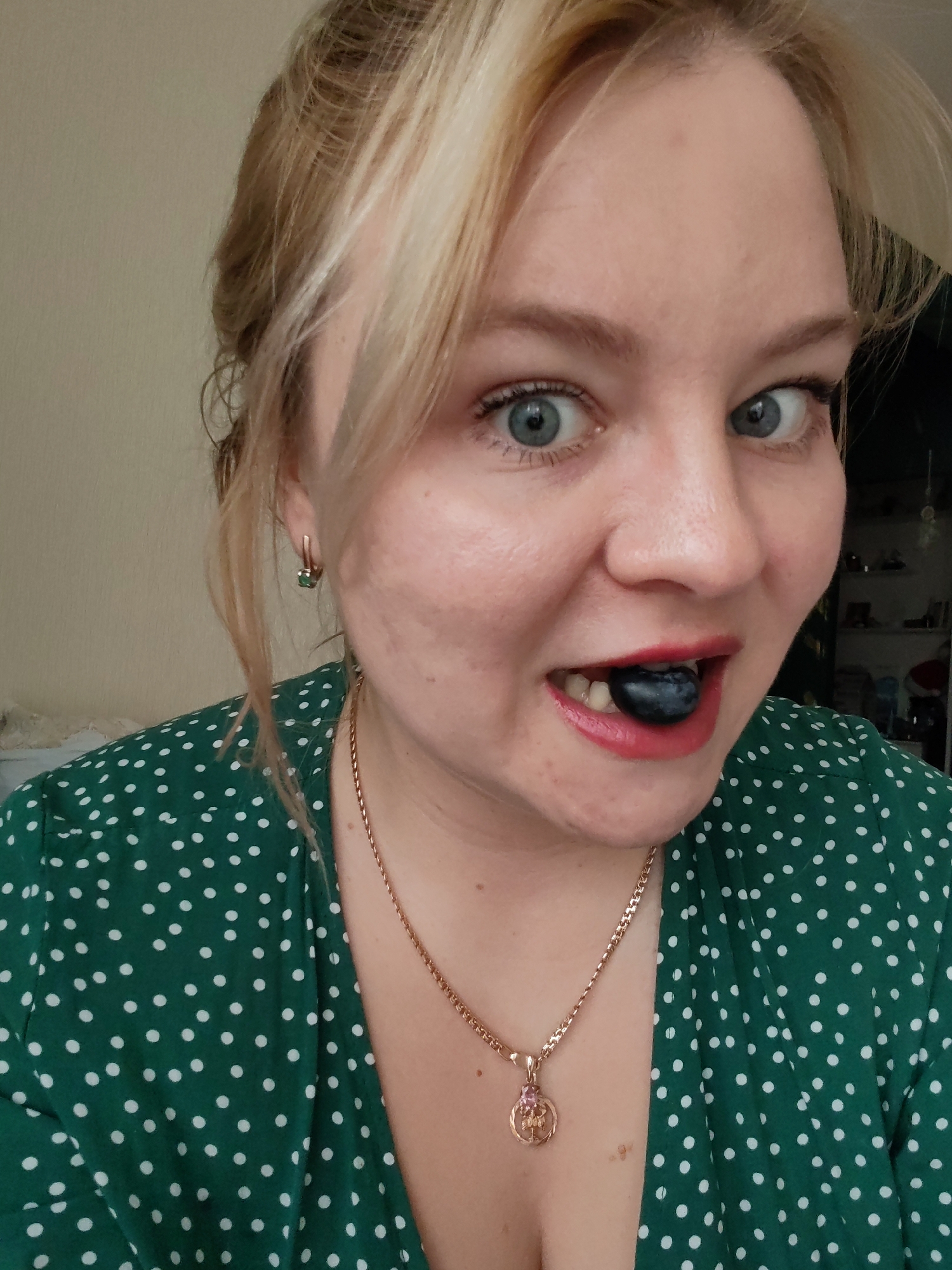 A woman with blonde hair, fair skin, and blue eyes is holding a large blueberry in her mouth. She is wearing a green dress with white polka dots and a gold necklace with a round pendant. She is looking directly at the camera with a slightly surprised expression. The background is slightly blurred, but appears to be an interior space with shelves and other objects visible. The location, Izhevsk, Russia, cannot be confirmed from the provided image.