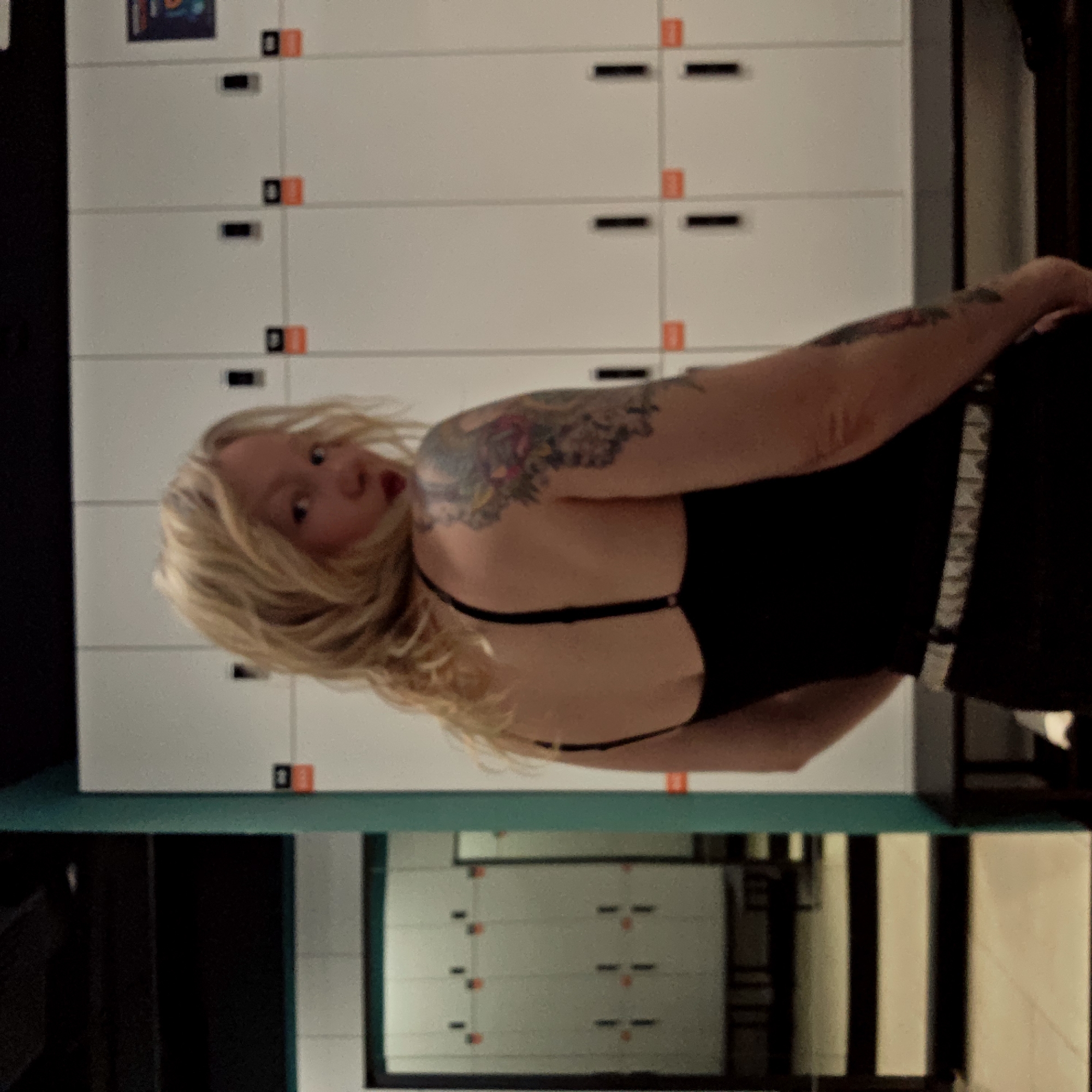 A woman with a tattoo on her left arm is standing in front of lockers, which are likely located in a gym or other athletic facility. The location is unknown; however, there is nothing to indicate that this is in Saint Petersburg, Russia. The lockers are white with black handles and orange markers. The woman is wearing a black top and a black skirt with a silver, studded belt. She is looking back over her shoulder. The background includes a reflection of the lockers and some black and white stripes, possibly indicating a hallway or another room.