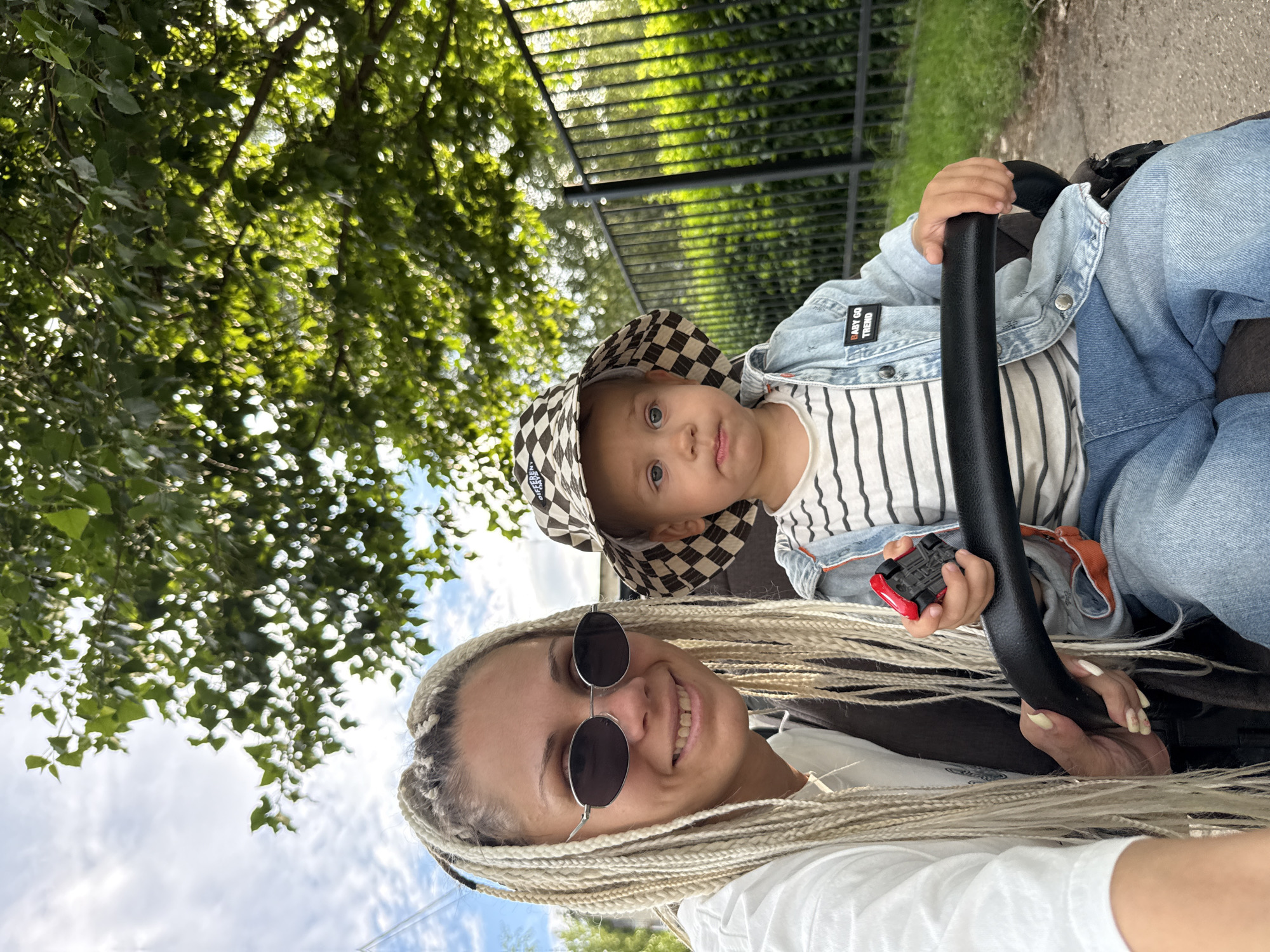 An adult female with blonde braided hair and sunglasses is taking a selfie with a young child in a stroller. The female is wearing a white t-shirt. The child is wearing a checkered bucket hat, a striped t-shirt, and a denim jacket with a patch that says "BABY GO TREND". The child is holding a small red toy car. They are outdoors, with trees and a fence in the background. The scene appears to be in a park or residential area, possibly in the city of Rusanovo, Russia, given the presence of greenery and a fence that resembles those found in Russian residential areas. The lighting suggests it is daytime.