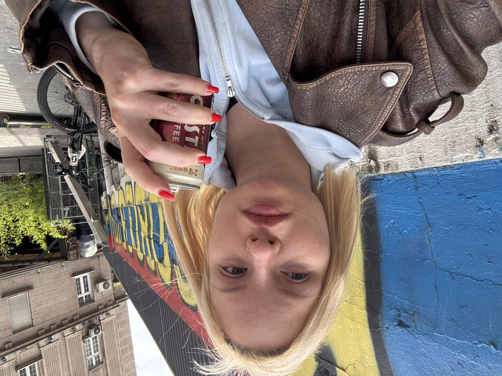 A medium shot of a person holding a can of iced coffee, taken from a low angle. The person has fair skin, blond hair, and is wearing a brown leather jacket over a light blue hooded sweatshirt. Their fingernails are painted red. The can is red and white, with text in English and Chinese.
The background includes an urban scene with a building visible on the left, painted with graffiti in blue, yellow, and red. In the distance, a bicycle is parked on the sidewalk. The image was taken in Belgrade, Serbia.