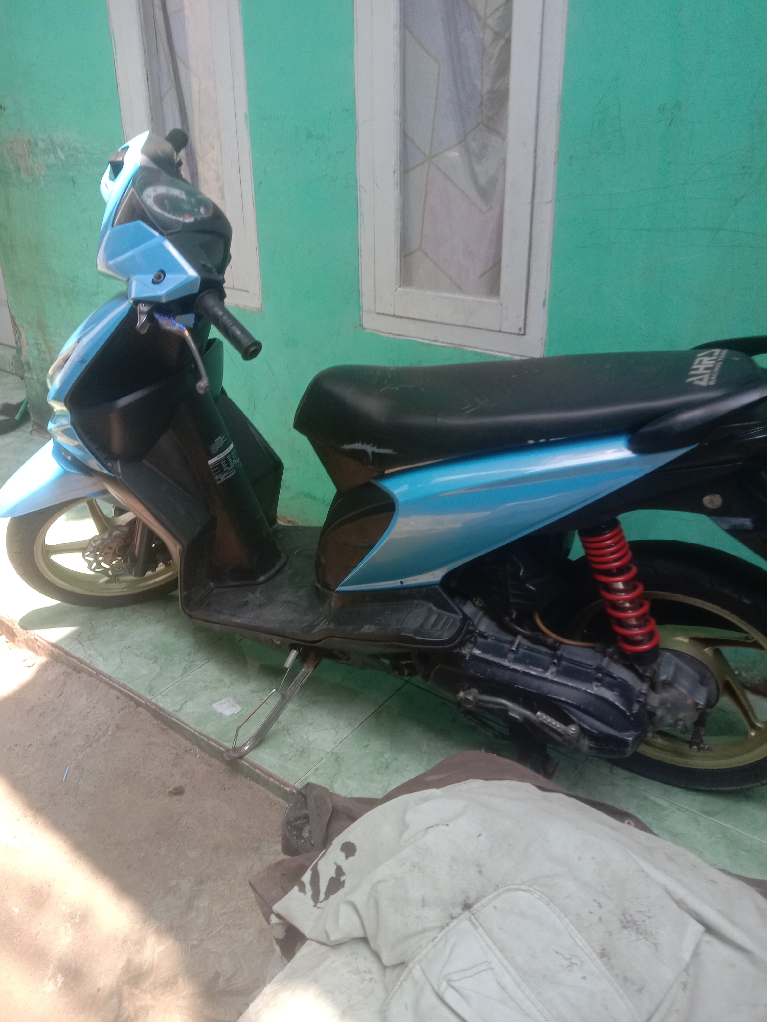 A light blue and black scooter is parked on a green tiled surface in front of a teal green wall with a white-framed window. The scooter's front wheel is visible on the left, with a disk brake. The handlebars and instrument panel are visible at the top left. The scooter has a black seat and a red rear shock absorber. To the right of the scooter, a white fabric object is partially visible on the ground, appearing to be a blanket or cover. The scene is outdoors and appears to be in a residential area.