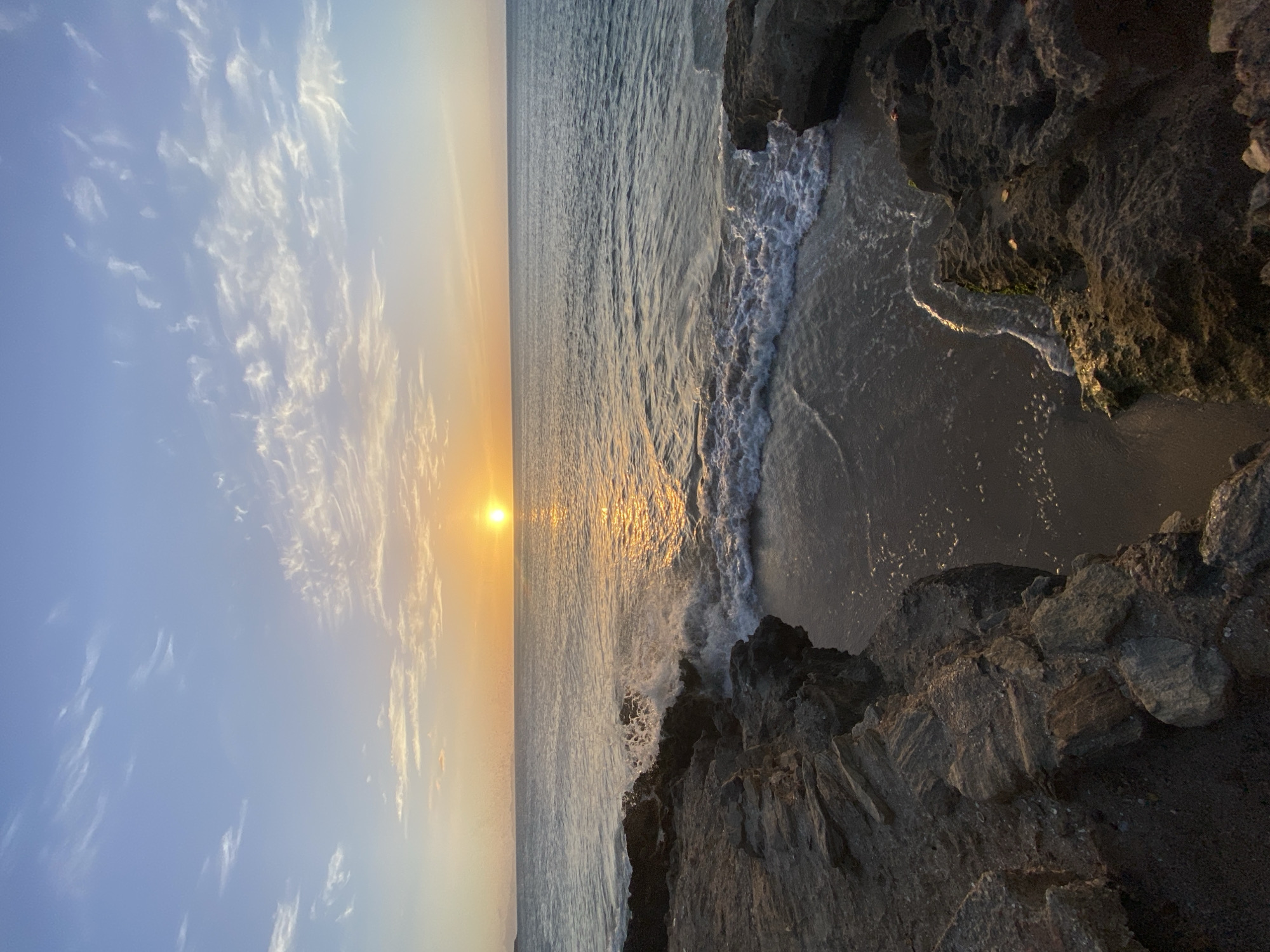 The image depicts a serene sunset scene at the coast of Juan Griego, Venezuela. The sun, a brilliant orb, is low on the horizon, casting a golden glow across the calm sea. The sky is a gradient of soft blues and oranges, with wispy white clouds scattered across it. Jagged rocks form the foreground, framing a small sandy cove where gentle waves lap the shore. The ocean is a deep blue, reflecting the warm hues of the setting sun. The overall impression is one of tranquility and natural beauty.