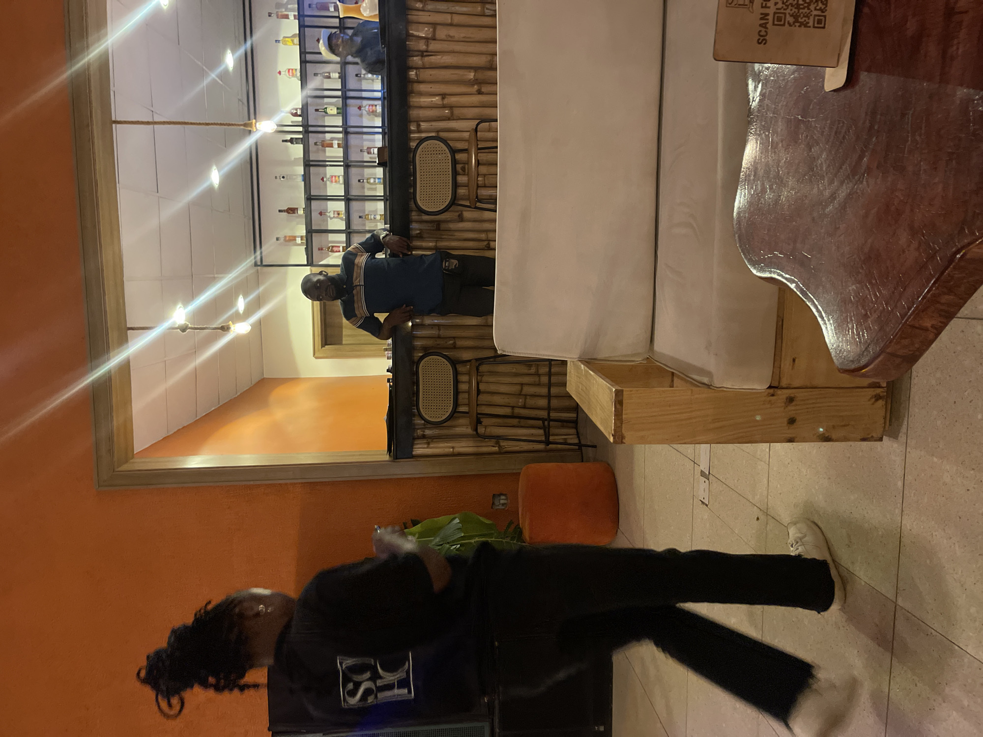 The image depicts an indoor bar scene, possibly in Gura Top, Nigeria, with warm orange walls and tiled flooring. In the foreground, a person with their back to the camera, wearing a dark hooded jacket with a white logo and black pants, is walking to the left. They have their hair in a braided updo. To the right, a beige couch made of wooden pallets and cushions faces a polished dark wood table. A QR code sign is placed on the table. Behind the couch, a bamboo-covered bar is visible with a bartender wearing a light-colored cap and a dark shirt, and another man in a striped polo shirt standing by the bar. Bottles are neatly arranged on shelves behind the bar. Two rattan-backed stools are positioned at the bar. An orange ottoman is visible near a large green plant. The lighting appears to be from overhead bulbs, some with visible ropes.