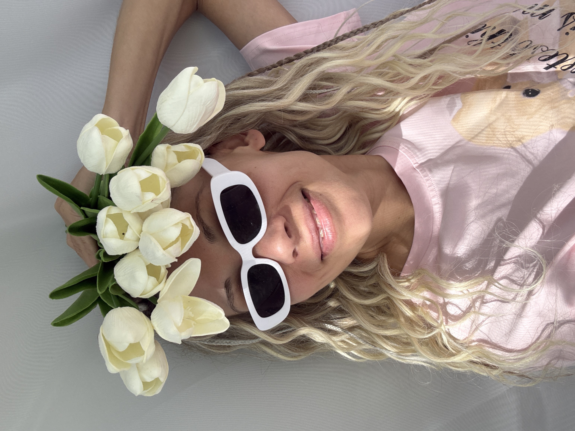 A woman is lying on a white surface, possibly a bed or soft ground. She is wearing a light pink shirt with an indiscernible graphic design. The woman has long, blonde hair with visible braids incorporated into it. She is wearing white-rimmed sunglasses with dark lenses. To her left, she holds a bouquet of white tulips with green leaves. Her arms are raised above her head. She is smiling with visible teeth. The photograph has a bright, natural lighting. It is impossible to confirm the location in Rusanovo, Russia, without further contextual information.