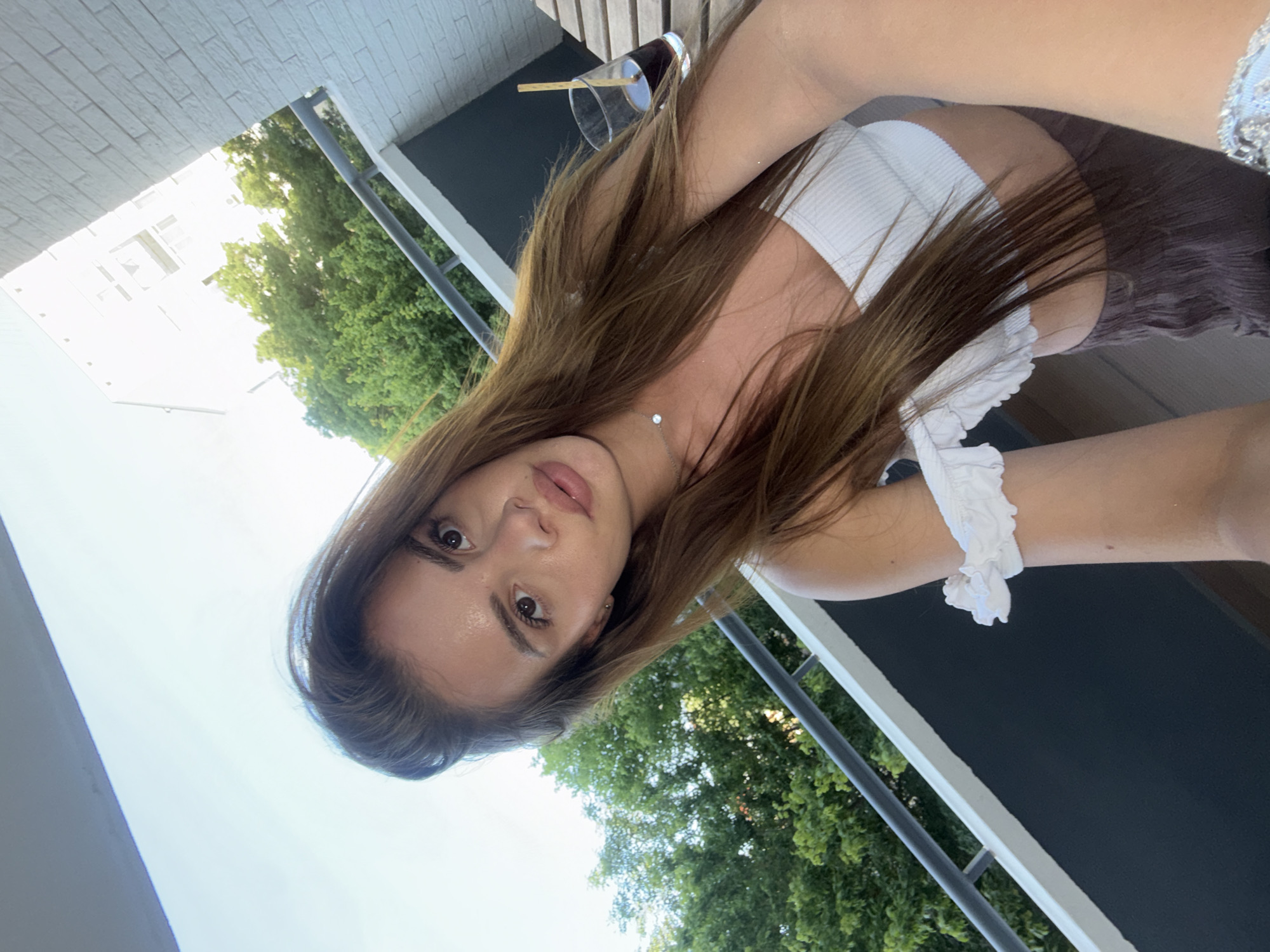 A young adult female is centered in a selfie taken outdoors on a balcony in Plovdiv, Bulgaria. She has long, dark brown hair, brown eyes, and wears light makeup with a pink lip color. Her attire consists of a white, ribbed, off-the-shoulder crop top featuring a ruffle detail on the left arm, and dark, textured fabric, likely trousers or a skirt, visible from the waist down. She wears a delicate silver necklace with a small, circular white pendant, and a metallic, possibly embellished, bracelet is partially visible on her right wrist. To her right, a glass containing a dark liquid and a light-colored straw rests on a slatted wooden surface. The balcony features a dark grey solid base, a white top edge, and a silver metal railing. Beyond the railing, lush green trees are visible. In the background, a multi-story building with a white brick-textured facade is prominent on the right, with a more distant, lighter-toned urban building visible further back. The sky above is bright and clear.