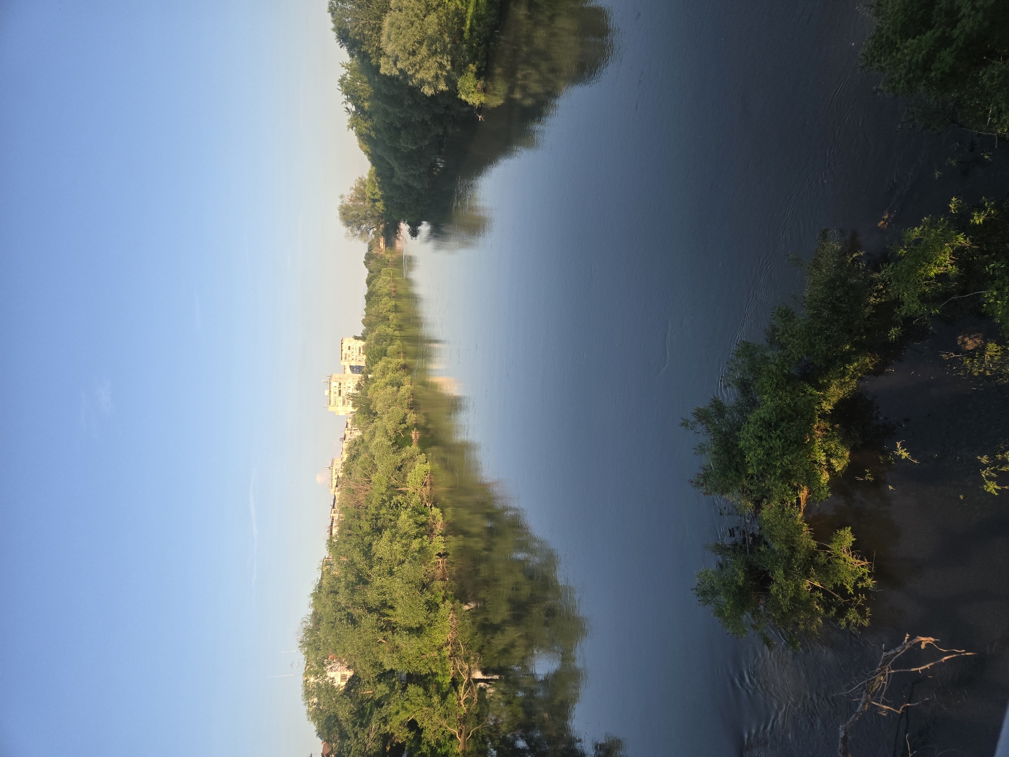 A landscape photograph depicts a calm body of water, likely a river or a large stream, framed by lush greenery on both banks under a clear sky. The water's surface reflects the sky and the vegetation, creating a mirror-like effect. The left bank features buildings interspersed with trees, suggesting an urban-natural integration. The right bank has dense vegetation that appears wilder and less manicured. The photograph is taken at a vertical orientation, which emphasizes the length and depth of the scene. The location, as specified, is the city of Unknown, Unknown.