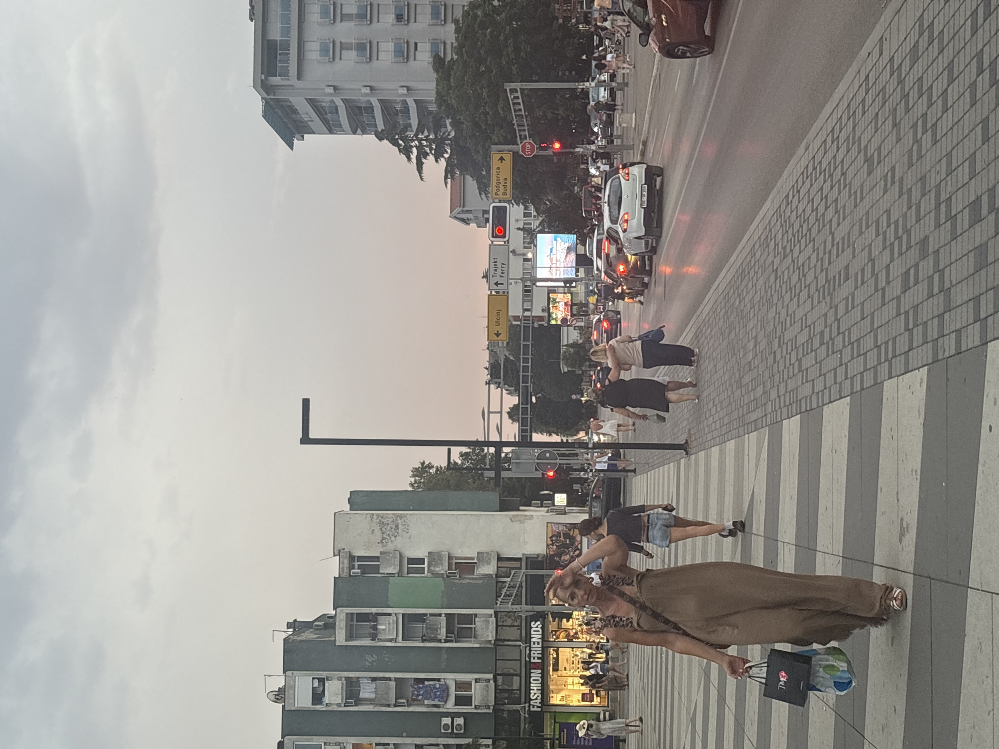 This is a street-level, full-frame, eye-level photograph taken outdoors on a cloudy day in the city of Bar, Montenegro. In the foreground, a woman with light-colored hair is walking towards the left of the frame. She is wearing a long, tan-colored dress and white sandals. She is carrying two shopping bags, one black with a red logo and the other a blue and white patterned bag. She has her right hand on her forehead, appearing to shield her eyes from the sun or heat.
Behind her, the sidewalk is paved with gray and white rectangular tiles in a striped pattern. Several other pedestrians are visible on the sidewalk and crossing the street in the background. Two women are walking away from the camera, one in a dark top and denim shorts, the other in a dark dress.
The street to the right of the sidewalk is busy with traffic. Several cars are driving, and some are stopped at a traffic light. The road surface is dark asphalt, and the traffic lights are red.
In the midground, there are several buildings. On the left, there is a tall, multi-story building with a dark green facade and white window frames. Signs for "FASHION & FRIENDS" are visible on the ground floor of this building, indicating a retail establishment. Further down the street, there are directional signs indicating "Ulcinj," "Trajekt Ferry," and "Podgorica Budva." A large digital billboard is also visible, displaying an image of a coastline.
On the right side of the street, a modern, multi-story building with a white facade and numerous balconies is prominent. Trees and other vegetation are interspersed between the buildings. The sky above is overcast with a pale, hazy light, suggesting either early morning or late afternoon. The overall impression is a bustling urban environment with pedestrian and vehicular activity.