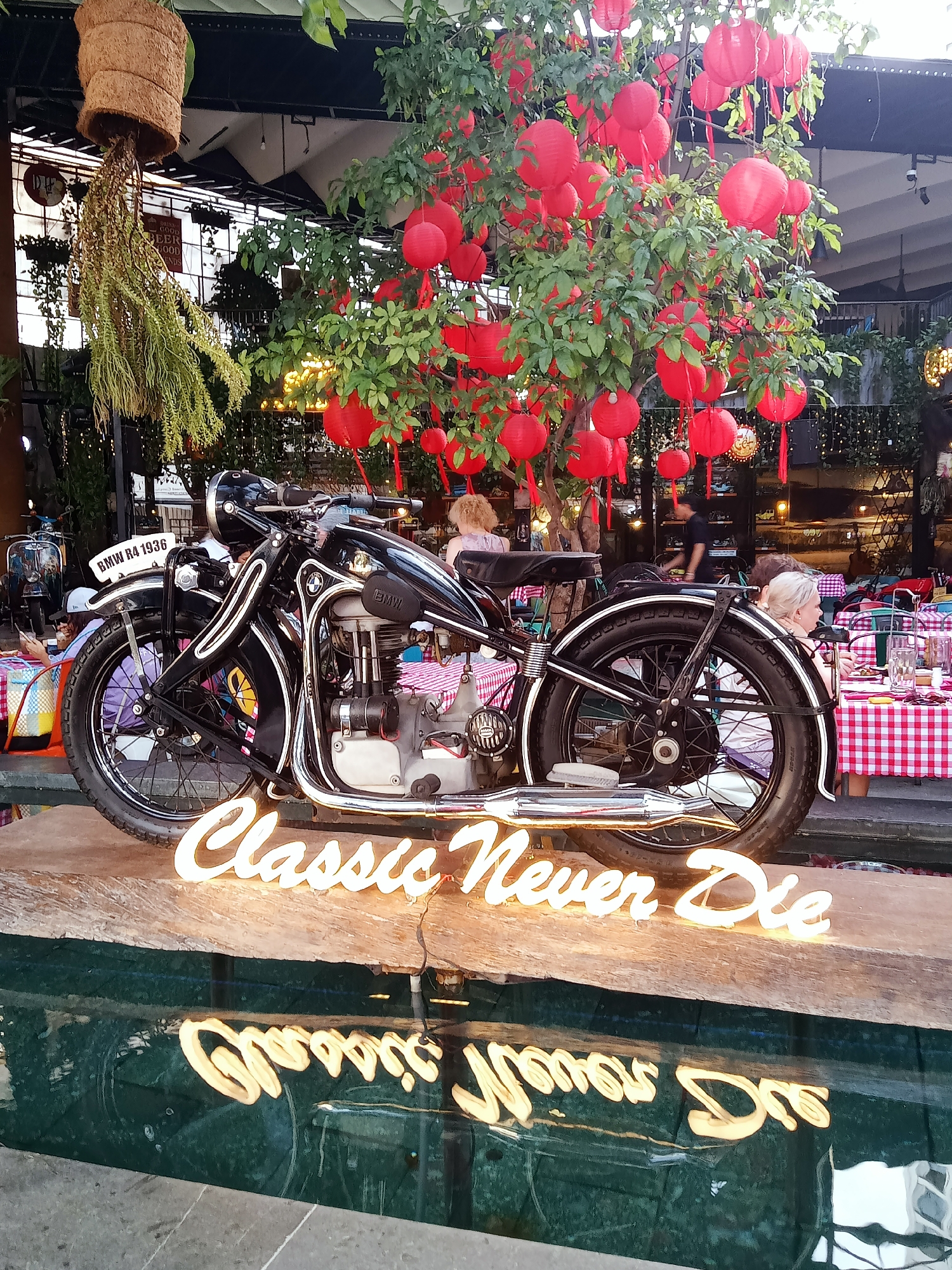 Captured in Nha Trang Ward, Vietnam, the image features a black BMW R4 1936 motorcycle displayed on a wooden platform. A neon sign reading "Classic Never Die" is positioned in front of the bike, its reflection visible in the water below. A tree adorned with red lanterns rises behind the motorcycle, adding a vibrant color contrast. The scene appears to be an outdoor dining area with people seated at tables covered in red and white checkered cloths. In the background, decorative plants hang from the ceiling of an open structure.
