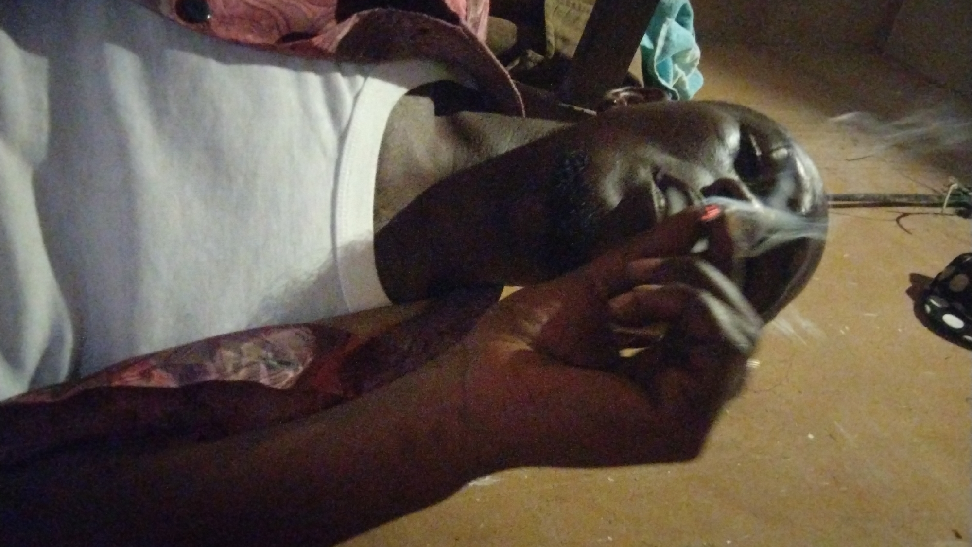 This dimly lit, close-up image captures a person smoking in Jos, Nigeria. The central subject, an individual with dark skin, is depicted from the chest upwards, wearing a plain white undershirt or tank top. Their head is tilted slightly back as they hold a lit cigarette or rolled tobacco product between their fingers, positioned near their mouth. Streams of smoke are clearly visible emanating from the cigarette and swirling around their face.
The person's expression appears calm and somewhat contemplative, though obscured by shadows and smoke. In the foreground, the hand holding the cigarette is prominent. The setting seems informal, with a light brown or tan-colored ground visible, possibly concrete or a simple floor. Hints of other objects are scattered in the background, including patterned fabric (likely clothing or a blanket), a folded light blue cloth, and a dark item with white polka dots on the far right. The low light levels suggest the photograph was taken at night or in an interior space with minimal illumination. There is no visible text in the image.