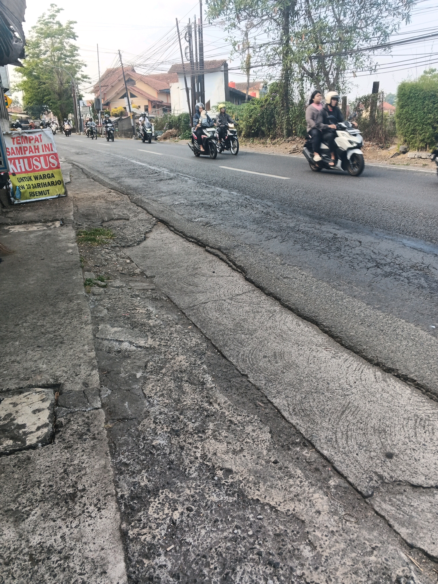The image depicts a street scene in Ungaran, Indonesia, likely on a paved road with a sidewalk. In the foreground, the sidewalk is made of weathered concrete, showing cracks and patches of wear. Adjacent to it, the road surface is also asphalt, with visible seams and textured patterns.
Several individuals are on motorcycles, traveling along the road. One white scooter carries two passengers, both wearing helmets, with the passenger in the back holding a bag. To the left, a yellow sign is visible on a stand, with text in Indonesian that translates to "This trash bin is specifically for residents of RW 10 Sariharjo Sisemut." Other motorcycles and riders are scattered further down the road.
The background shows buildings with tiled roofs and trees lining the street. Overhead, a complex network of electrical wires crisscrosses the sky. The overall impression is a typical, bustling urban or semi-urban street in Indonesia. The lighting suggests it is daytime, possibly late afternoon or early morning given the relatively soft light.