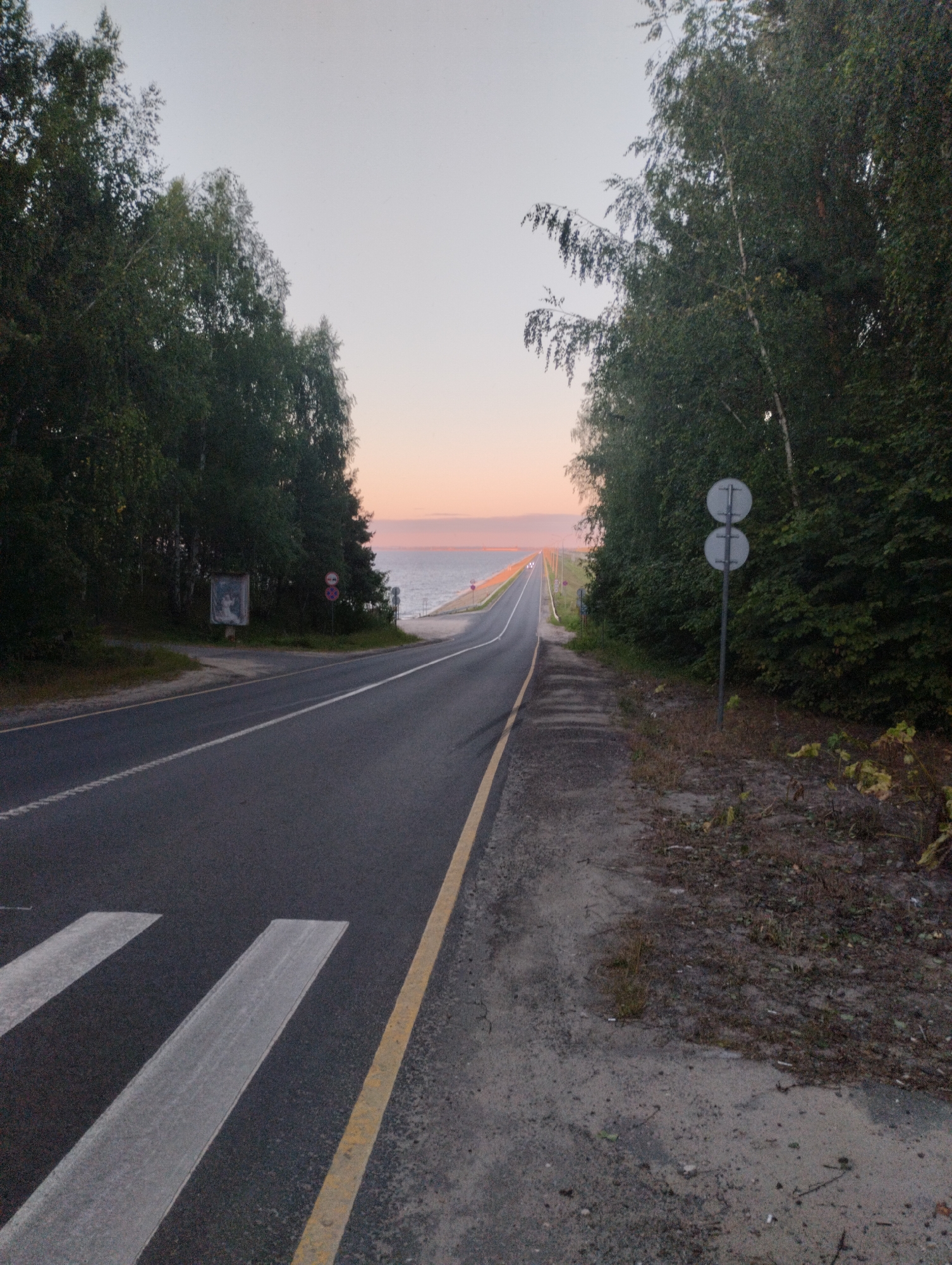 The image depicts a road in Zavolzhye, Russia, at dusk. A paved road with white lane markings runs through a forested area. On the left side of the road, there are trees and a signpost with a framed picture. On the right side of the road, there are more trees and a double round signpost. The road leads towards a body of water, possibly a river or lake, in the distance, with a faint orange glow on the horizon indicating sunset. The sky is a pale gradient of gray and pink. There are no people visible in the image.