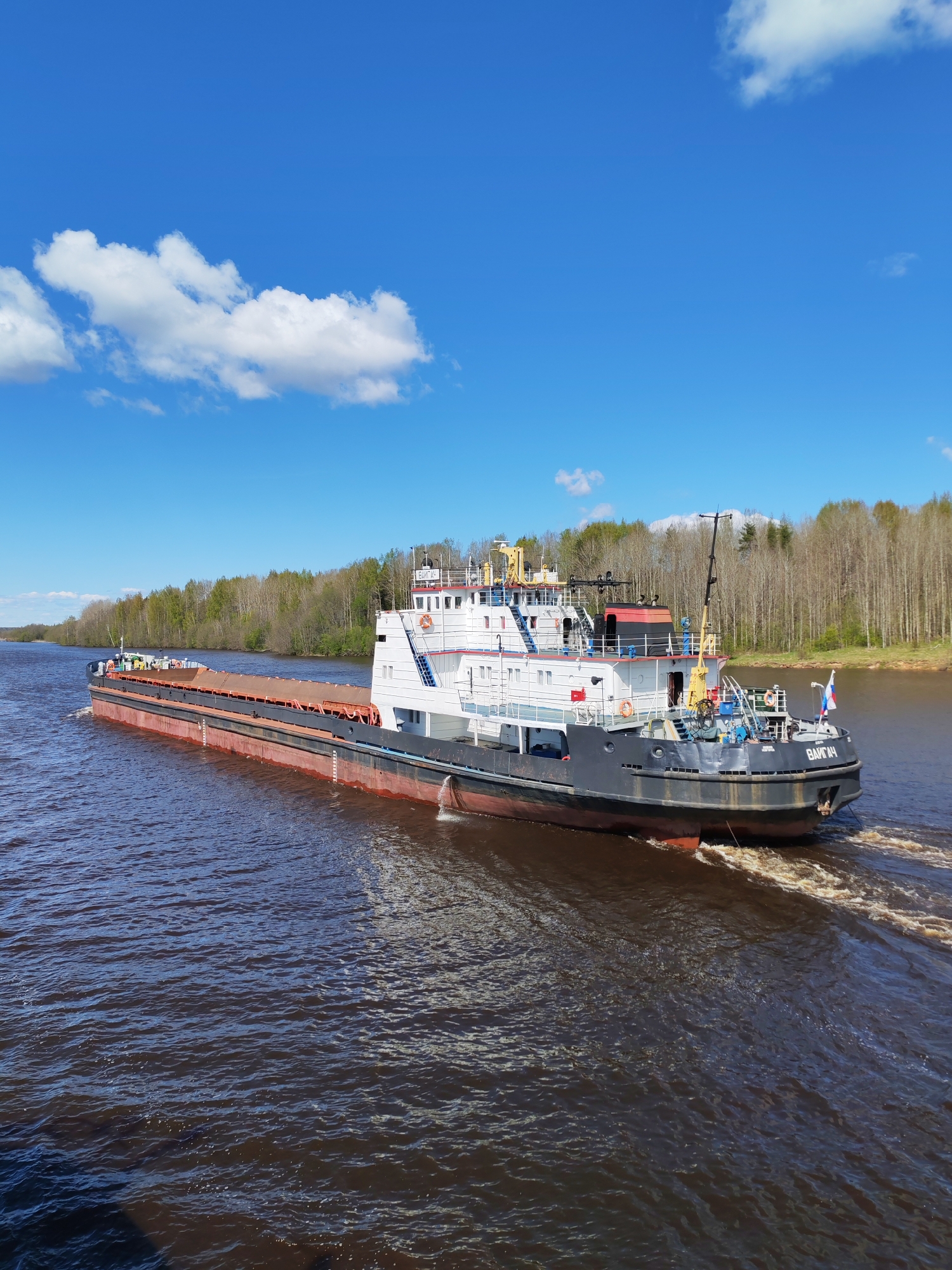 The image depicts a large barge traveling on a river. The barge is primarily grey with white and red accents. It has a multi-level superstructure. The river is a dark brown color, and the barge is creating a wake as it moves through the water. On either side of the river are dense forests with bare trees. The sky is blue with scattered white clouds. The barge is in Pruzhinino, Russia.