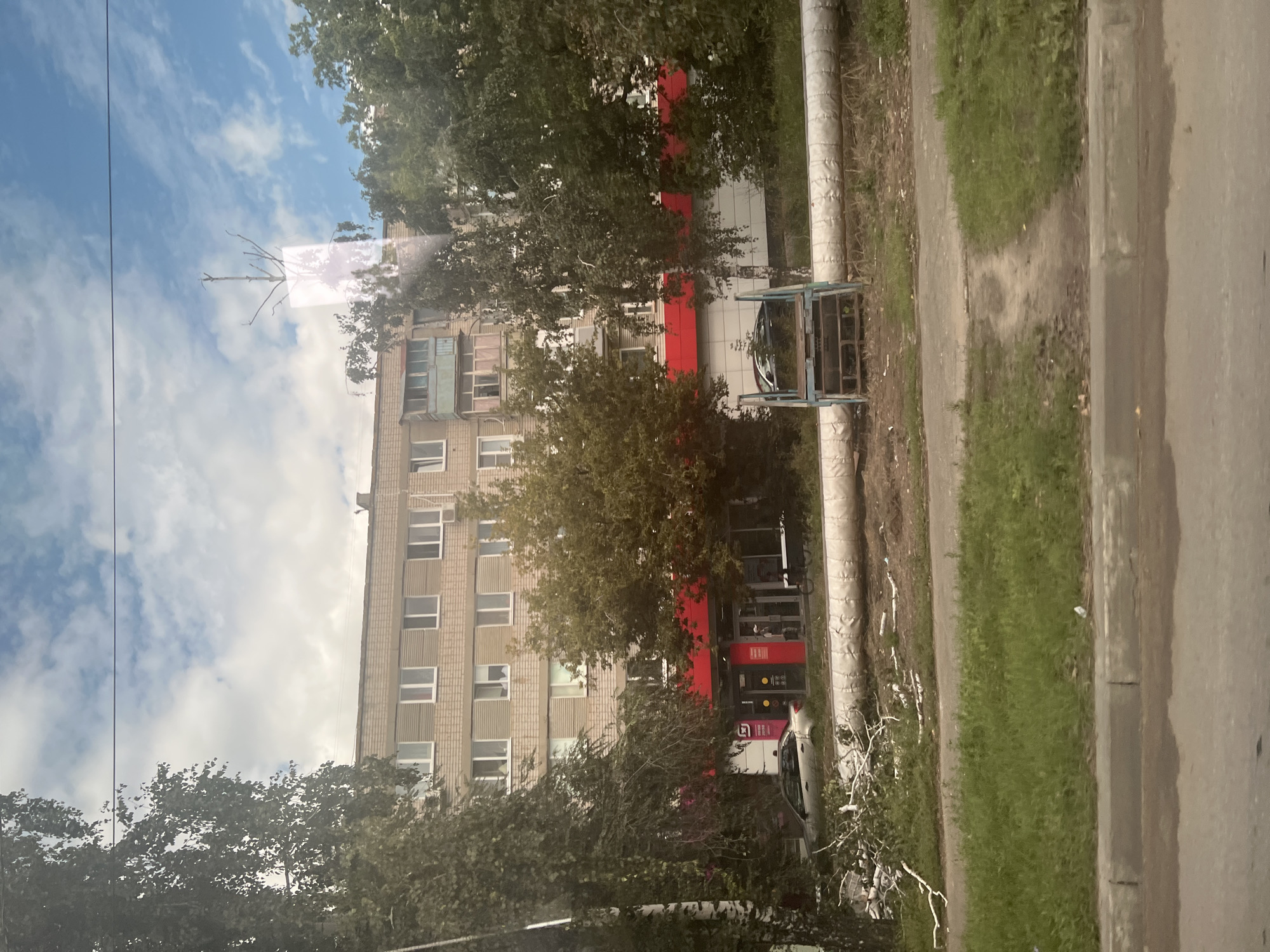 The image depicts an outdoor scene in a city, likely Ulyanovsk, Russia, focusing on a multi-story apartment building and its immediate surroundings. The building, constructed with light-colored bricks, has multiple floors with uniformly spaced windows, some of which are open or have visible contents. A balcony with an enclosed section is visible on one of the upper floors. The ground floor of the building features a commercial establishment with a prominent red facade, large windows, and signage. A silver car is parked in front of this establishment, and a person can be seen standing near the entrance.
In front of the building, a large, insulated pipe runs horizontally across the frame. Adjacent to this pipe, there is a small, blue, metal structure that appears to be a set of stairs or a platform. To the left of the pipe, a pile of fallen branches and debris, possibly from a tree, is visible on the grassy area. Several trees with green foliage are present in the foreground and between the viewer and the building, partially obscuring the view of the apartment complex. The sky above is blue with scattered white clouds, suggesting daytime. A dirt path with some worn areas leads through a grassy area in the foreground. The bottom portion of the image shows a section of a paved road with a curb and some wet patches, indicating a street or sidewalk area.